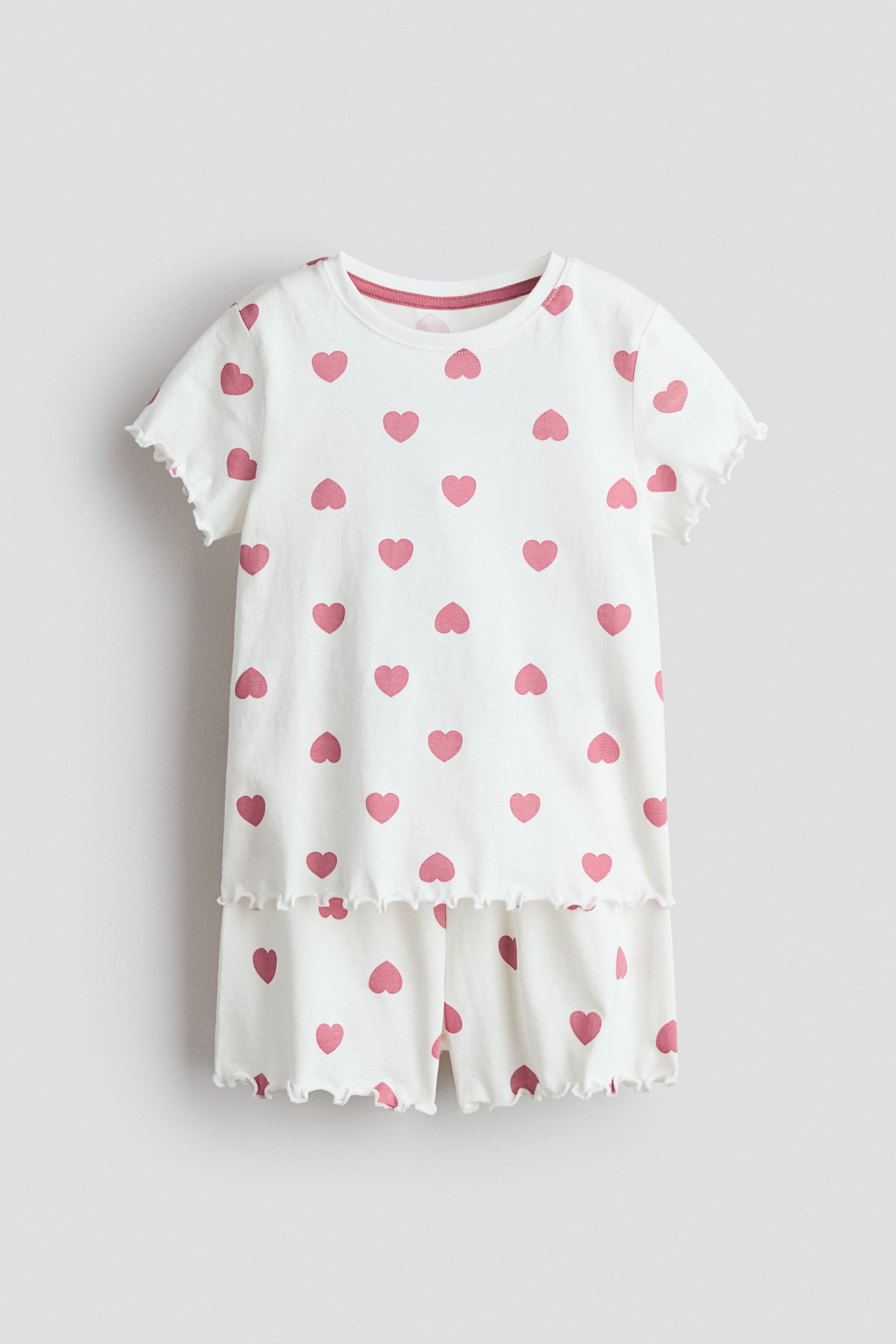View larger image: A set of white jersey pyjamas, presented FRONT, features an all-over pink heart print. The short-sleeved top displays a round, pink rib-trimmed neckline and overlocked, wavy edges at the cuffs and hem, while the matching shorts also show overlocked, wavy edges at the hems.