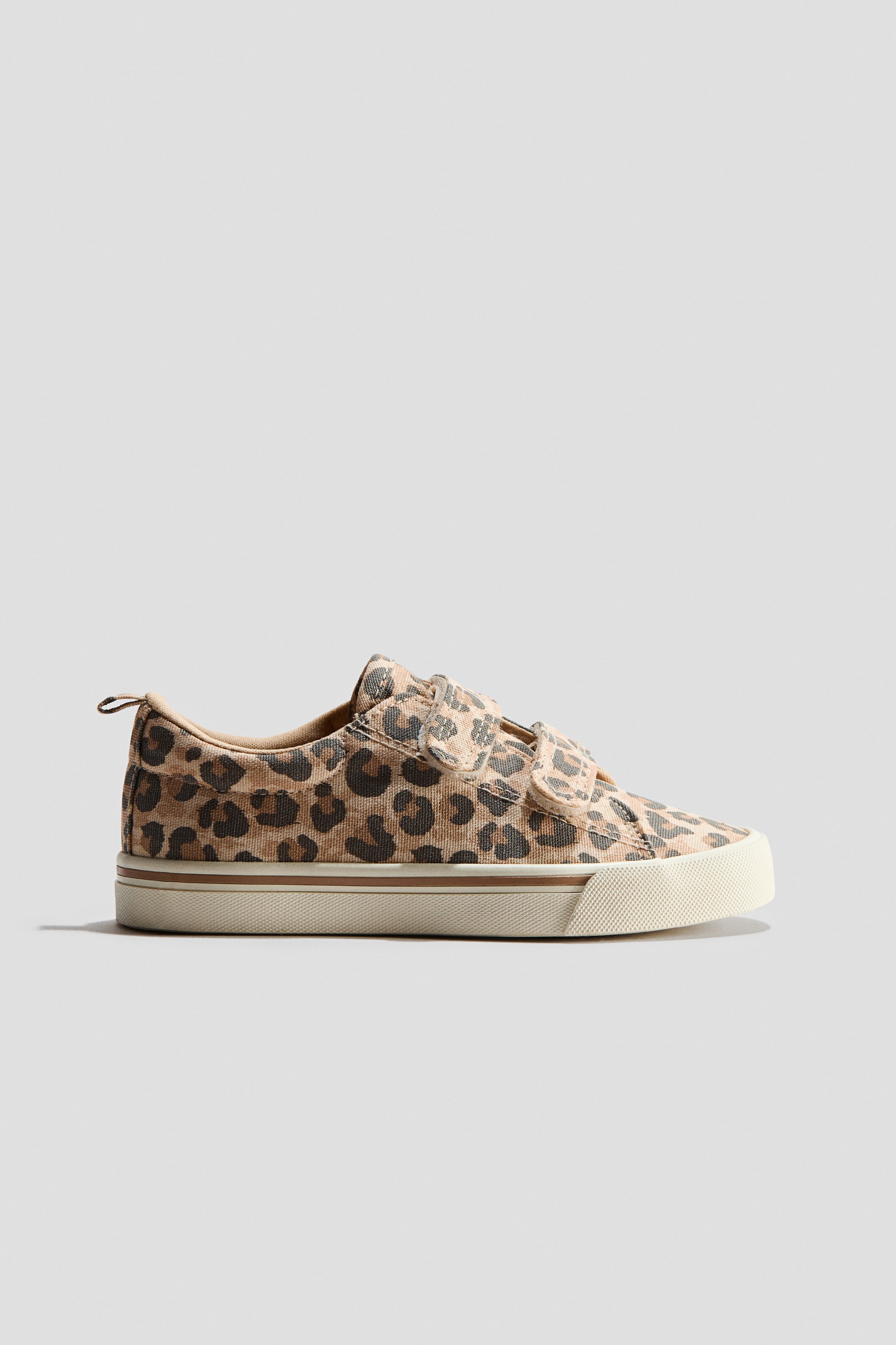 View larger image: This trainer features a leopard print canvas upper with two Velcro straps securing the front. It showcases an off-white rubber sole with a textured toe cap and a thin brown stripe detailing the side.