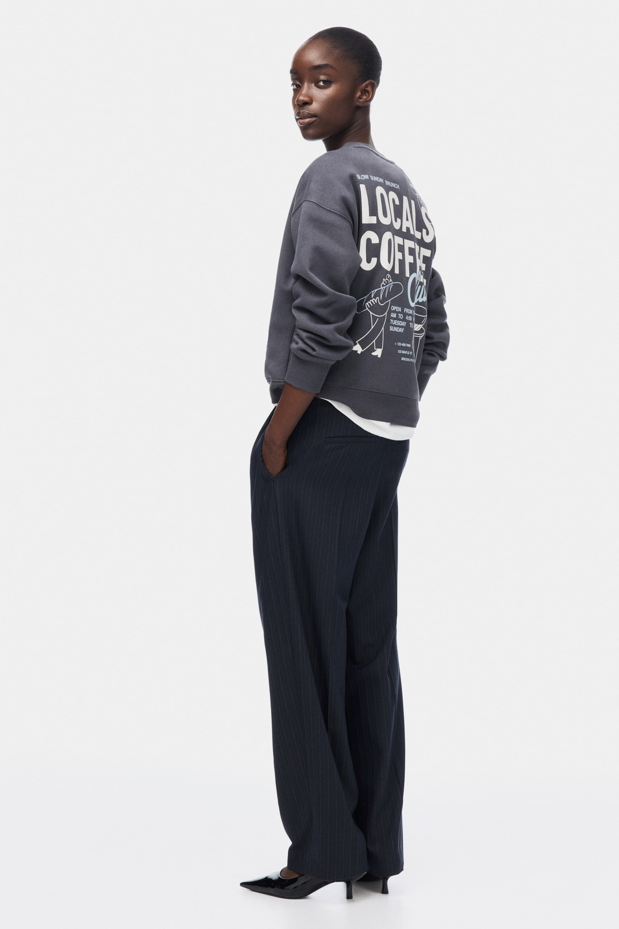 View larger image: A grey crewneck sweatshirt is depicted from a back-right perspective, showcasing a prominent white graphic on the back with the text "LOCALS COFFEE," accompanied by smaller text details and an illustration of a figure holding bread. It exhibits a relaxed fit, with a white shirt layered beneath its ribbed hem.