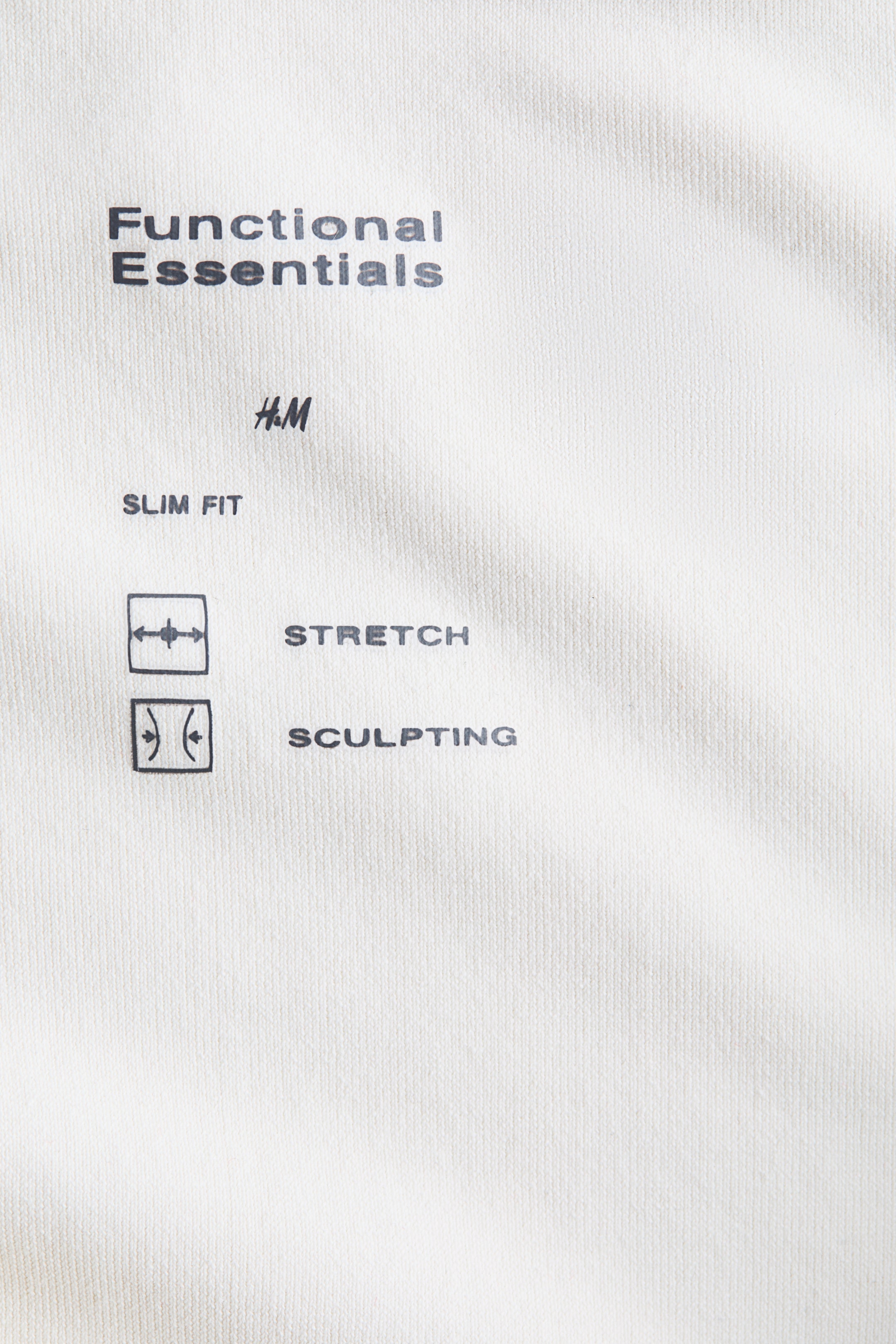 Büyük resmi görüntüle: Beyaz bir kumaş, siyah metin ve simgelerle donatılmış. Kumaşın üzerinde "Functional Essentials," "H.M," "SLIM FIT," "STRETCH," ve "SCULPTING" yazıları basılı. "STRETCH" ile dışarı doğru yönelen oklarla bir kare simge ve "SCULPTING" ile içe doğru kıvrılan oklarla bir kare simge eşlik ediyor.