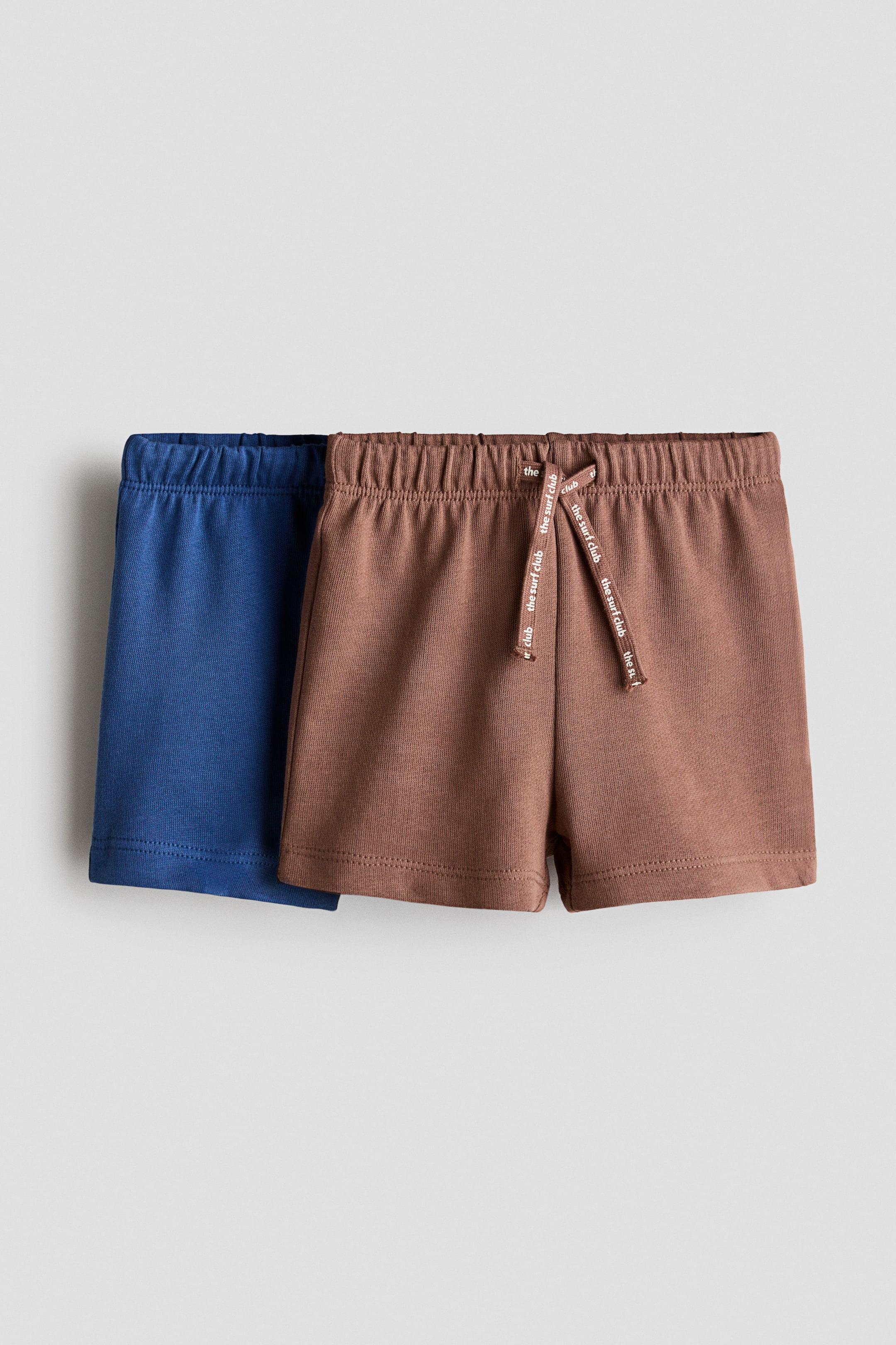 View larger image: The shorts are displayed as a set of two, one in blue and the other in brown, both crafted from soft, lightweight cotton sweatshirt fabric with an elastic waistband. The brown shorts feature a drawstring, adorned with the repeated text "the surf club," tied in a bow.