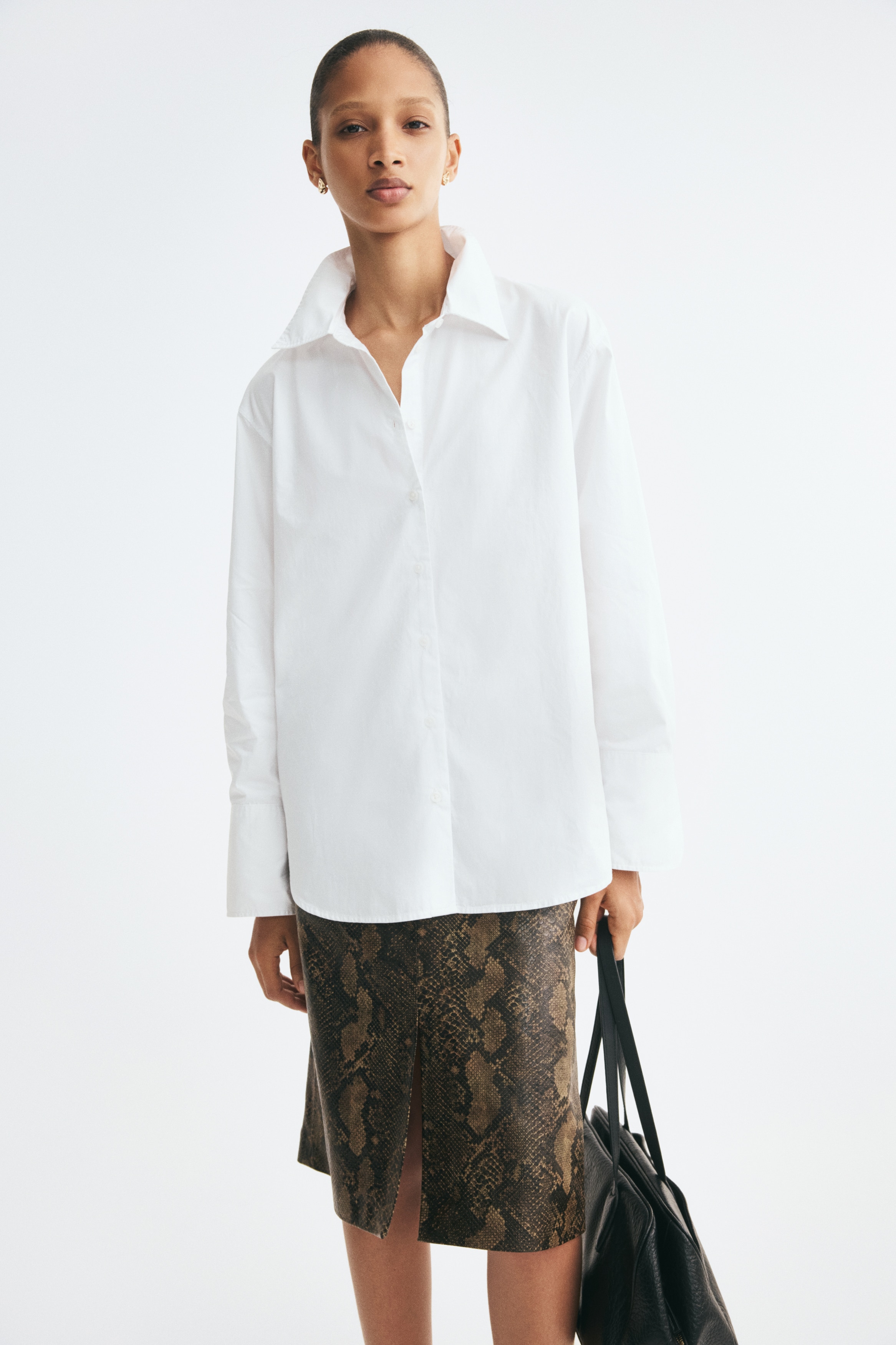 Mặt TRƯỚC của chiếc áo sơ mi dáng rộng làm từ vải poplin cotton sắc nét có cổ áo cổ điển và hàng nút cài toàn bộ phía trước. Áo có thiết kế vai rơi nhẹ nhàng, tay áo dài với cổ tay áo rộng có nút cài, và gấu áo cắt thẳng với đường xẻ tròn hai bên.