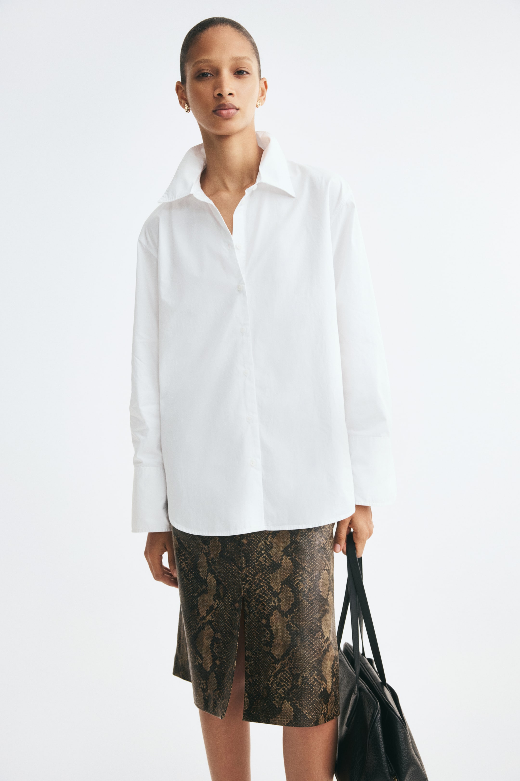 Voir une image plus grande: Une chemise blanche surdimensionnée en popeline de coton impeccable est portée de face, avec un col classique, une fermeture boutonnée sur le devant et des épaules légèrement tombantes. La chemise met en valeur de longues manches avec de larges poignets boutonnés et est portée non rentrée sur une jupe marron à motif peau de serpent.