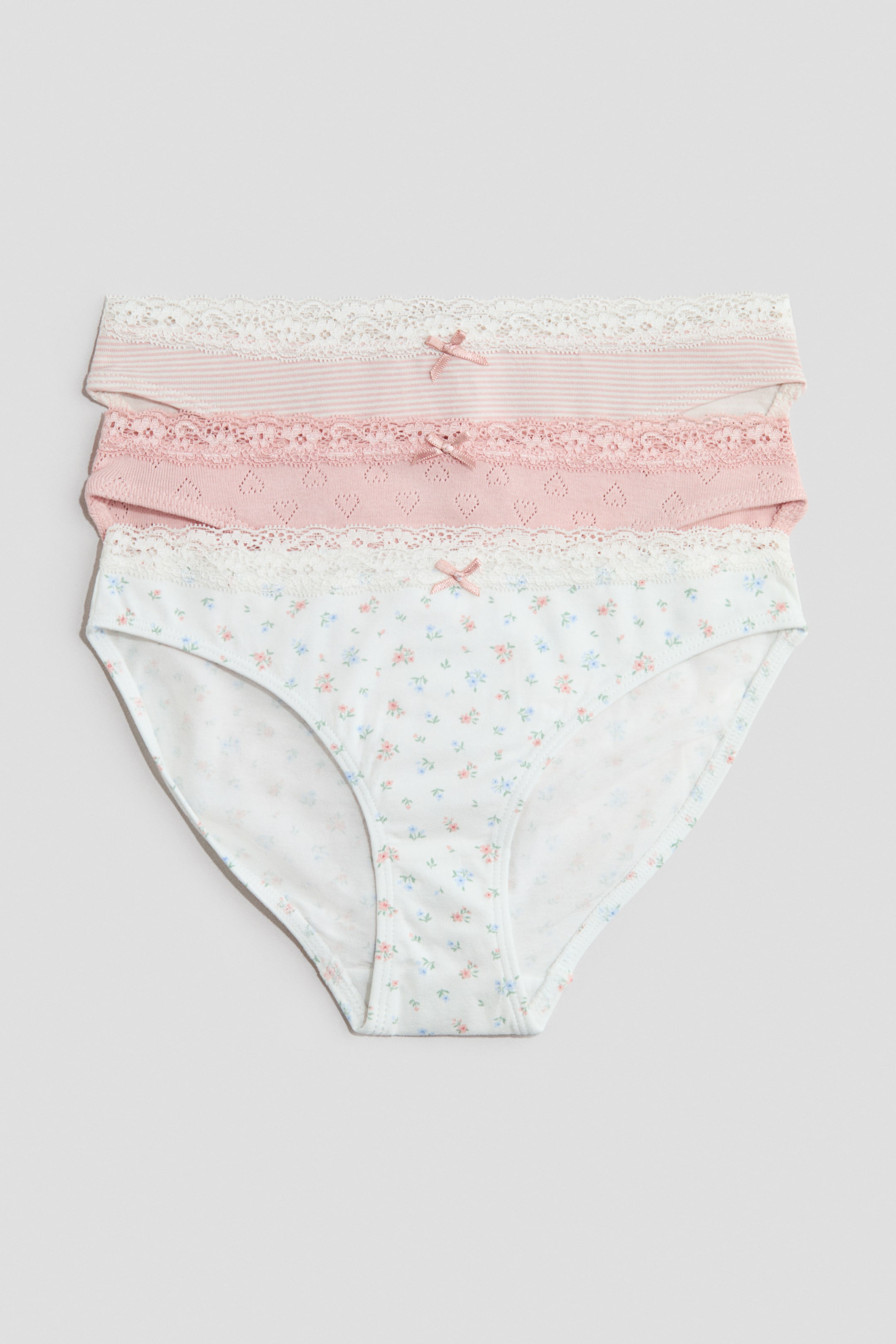 View larger image: Three soft cotton jersey briefs are stacked, presenting their front. The top brief features light pink and white stripes, the middle brief showcases a solid light pink with a heart-shaped eyelet pattern, and the bottom brief displays a white base with a small floral print; each is detailed with an elasticated waist, adorned with white lace trim and a small pink satin bow at the centre front.