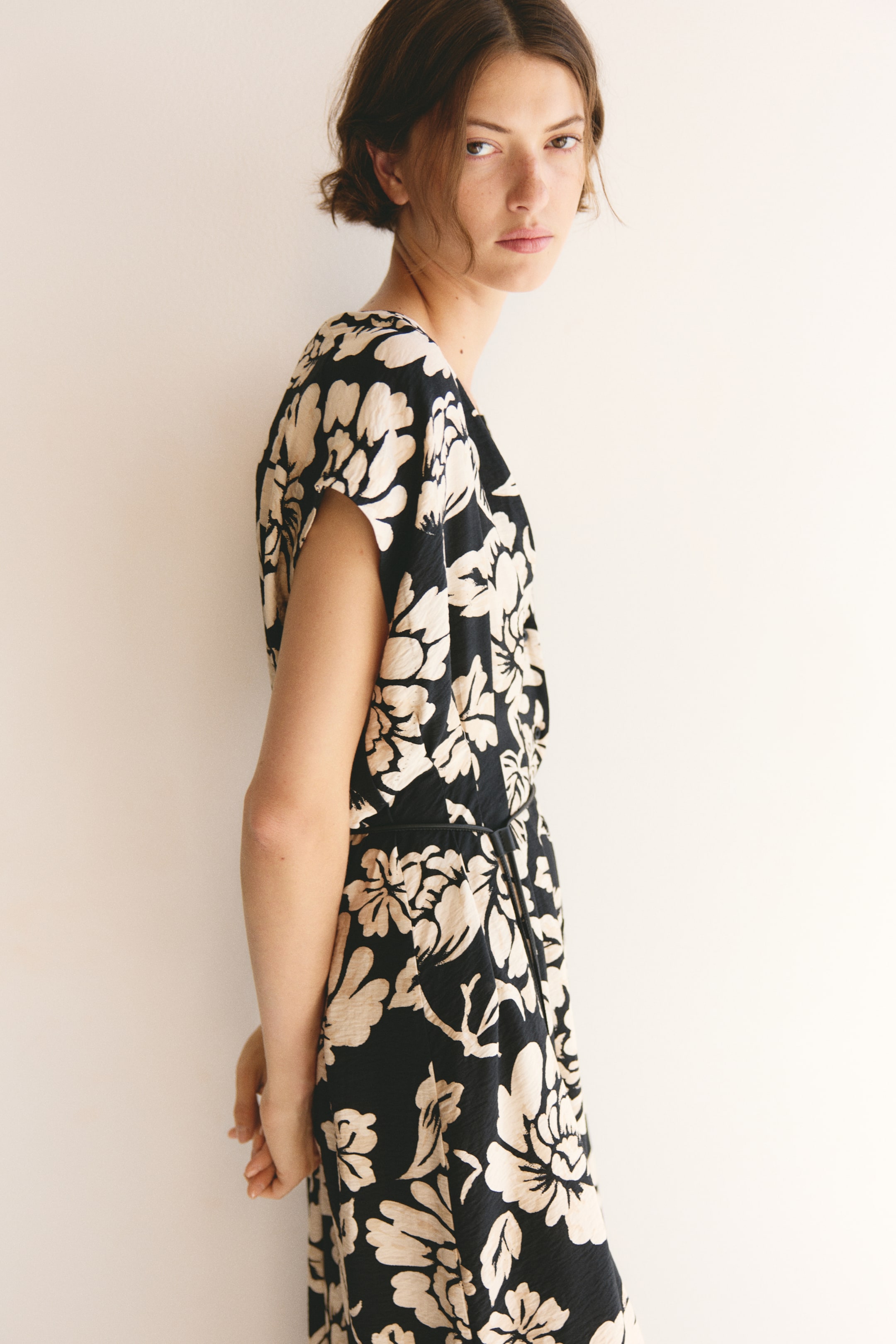 Ver imagem maior: Um vestido com fundo preto e padrão floral leve, com mangas curtas e cintura ajustada com um cinto fino preto, está a ser usado pela modelo. A modelo está de pé com as mãos entrelaçadas atrás das costas.
