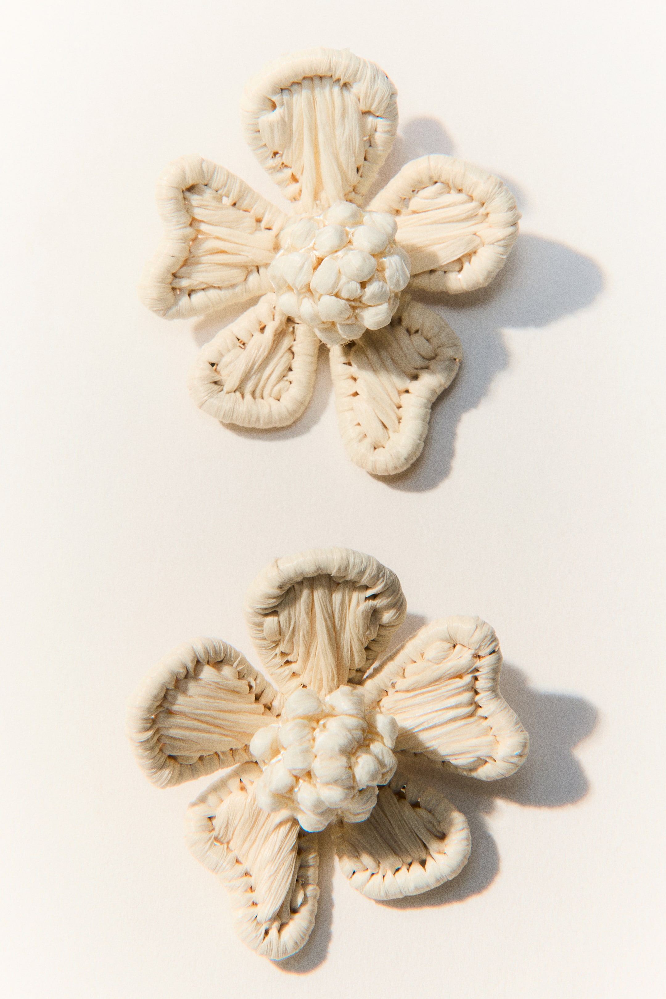 View larger image: Two cream-coloured decorative flowers, crafted from woven natural fibres, are lying flat and are positioned vertically. Each flower comprises five petals with thick, wrapped borders and textured interiors, while a central cluster of small, rounded elements forms its core.