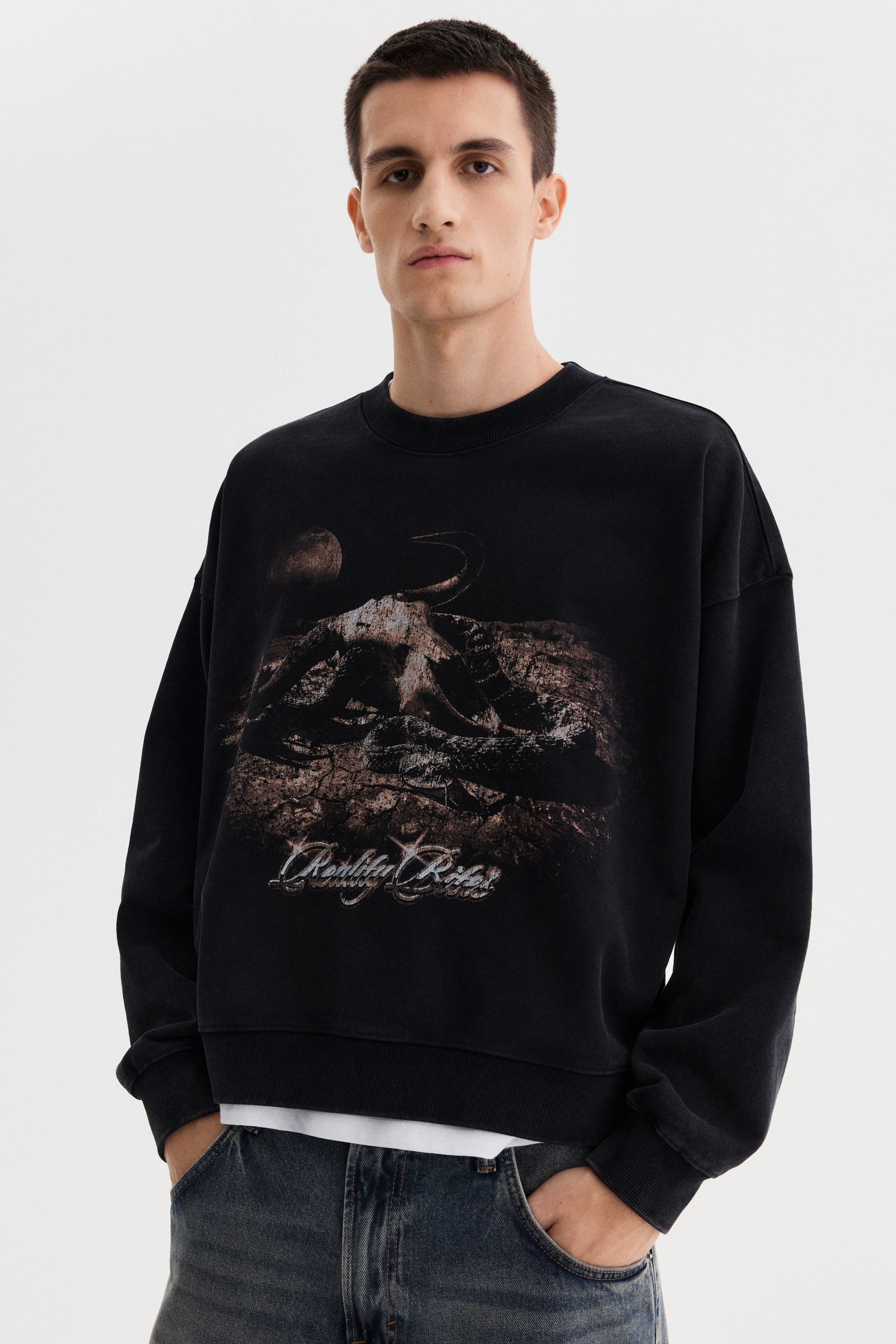 View larger image: A black crewneck sweatshirt features an oversized fit with ribbed cuffs and hem, and a large graphic print on the front. The print shows a textured, horned skull on cracked ground beneath a moon, with the text 'Reality Rider' below it. A white T-shirt is visibly layered under the hem.