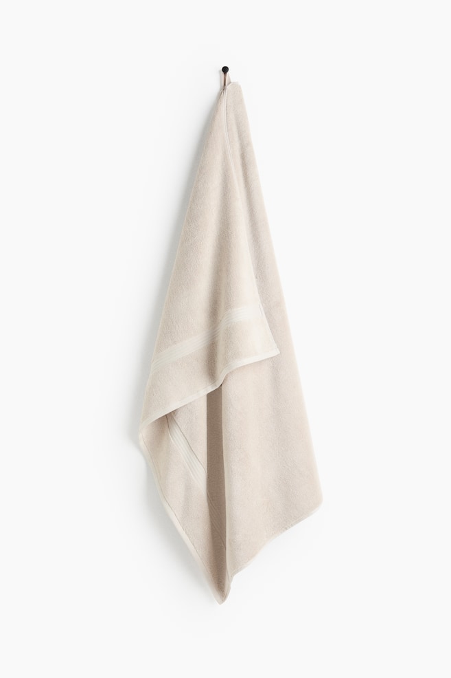 A light beige towel is hanging from a hook, showcasing a soft texture and a subtle horizontal decorative band. It drapes with gentle folds, with one section layering over another. - 1