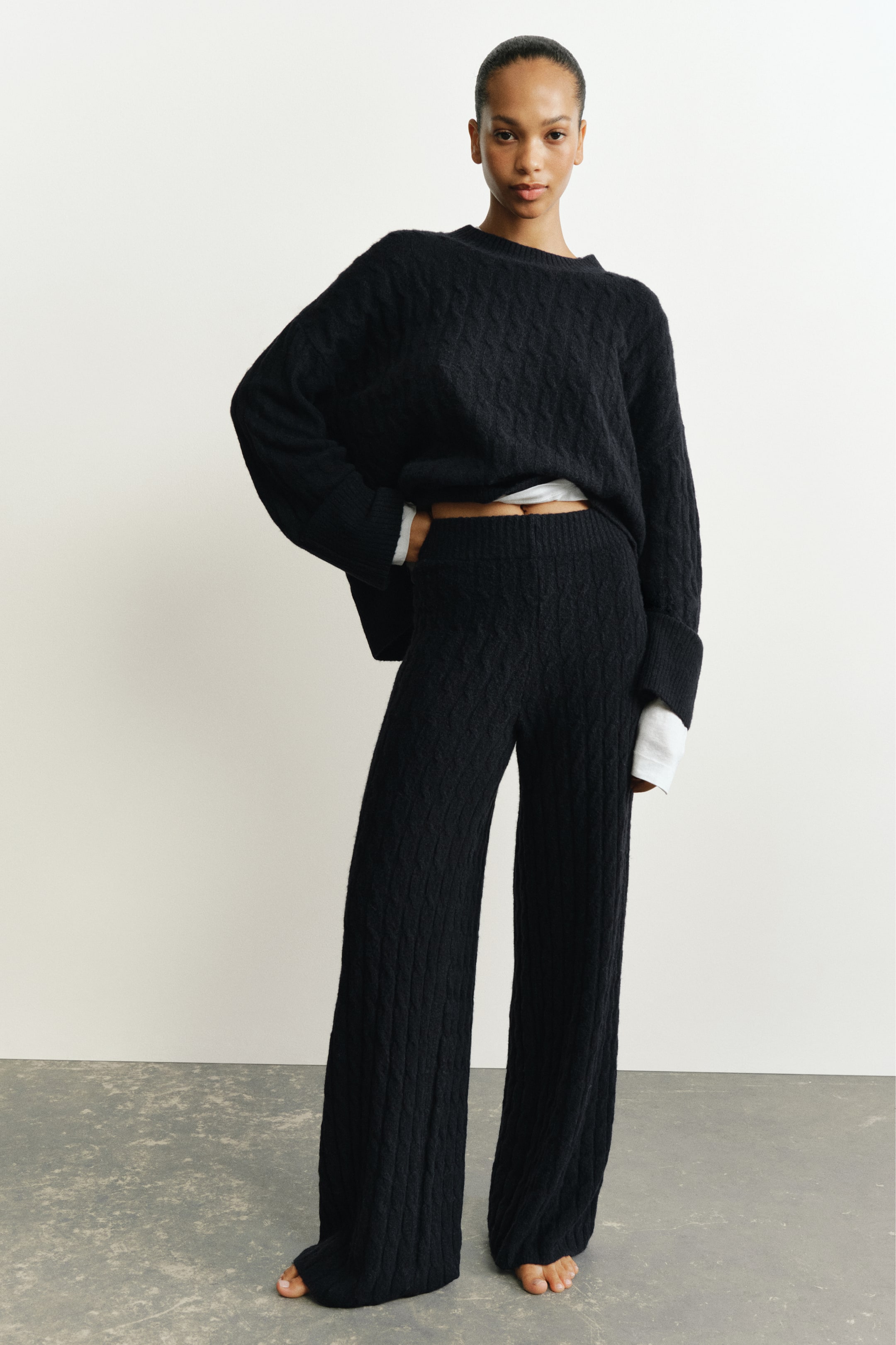 Se større billede: Sorte kabelstrikkede bukser med ribbet talje og afslappede, lige ben bæres af modellen. Bukserne er stylet med en matchende sort kabelstrikket sweater, lagt over en hvid langærmet top.
