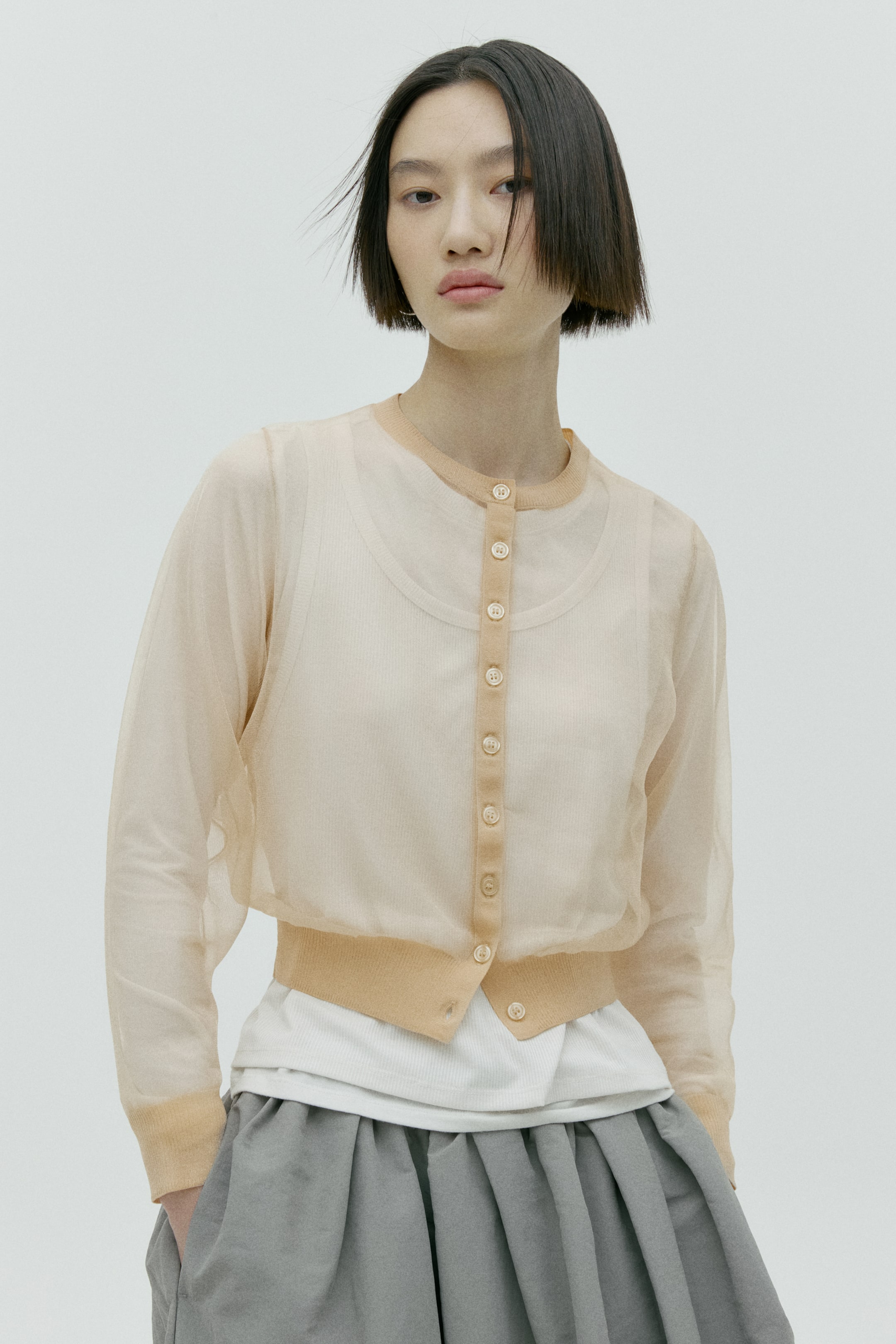 이미지 확대 보기: 연한 피치색 시어 가디건은 라운드 네크라인과 전면 전체 버튼 플래킷이 특징입니다. 리브드 커프스가 있는 긴 소매와 리브드 처리된 밑단을 갖추고 있으며, 흰색 리브 탑 위에 레이어드되어 있습니다.