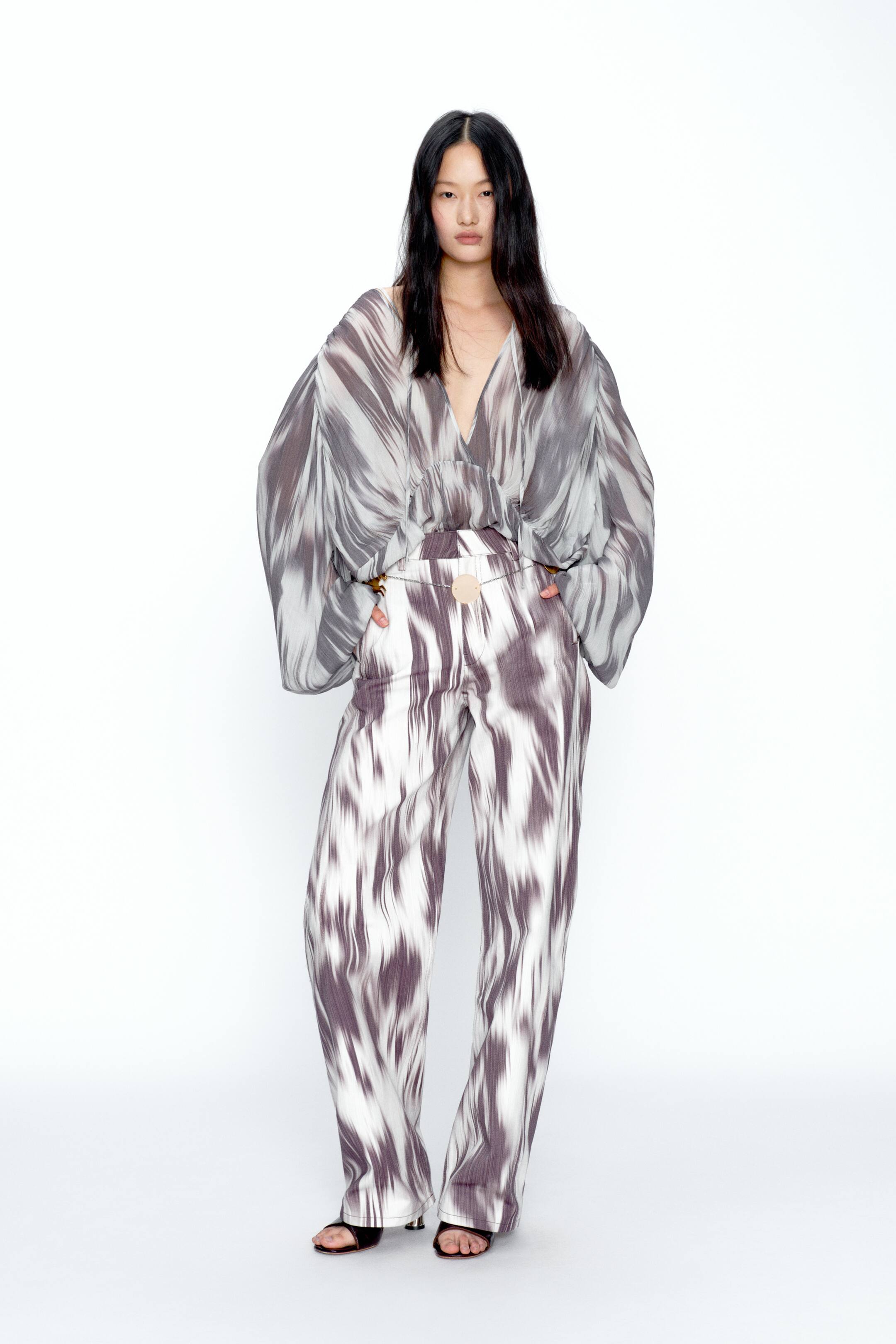 View larger image: A coordinated two-piece set featuring a fluid V-neck top and wide-leg trousers, both in an off-white and muted purple-grey abstract pattern. The top is softly gathered at the waist, while the high-waisted trousers are styled with a slim chain belt finished with a circular buckle.