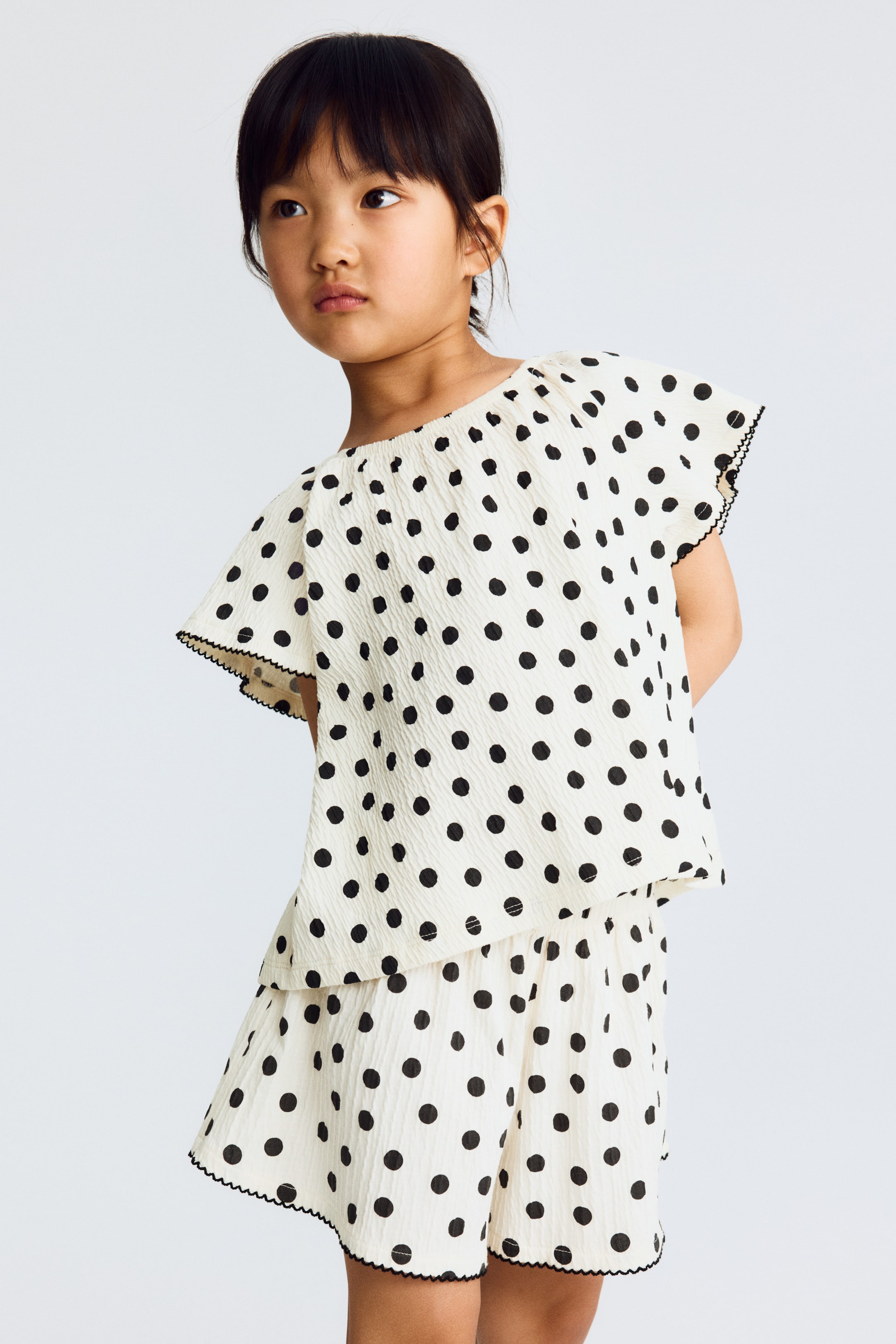 Grotere afbeelding bekijken: Een tweedelig kinderkledingstuk, licht naar rechts gericht, is gemaakt van een gebroken witte gekreukelde stof met zwarte stippen. De top is ontworpen met een elastische off-the-shoulder halslijn en korte fluttermouwen, terwijl de bijpassende shorts de set compleet maken, beide stukken zijn afgewerkt met een zwarte geschulpte rand.