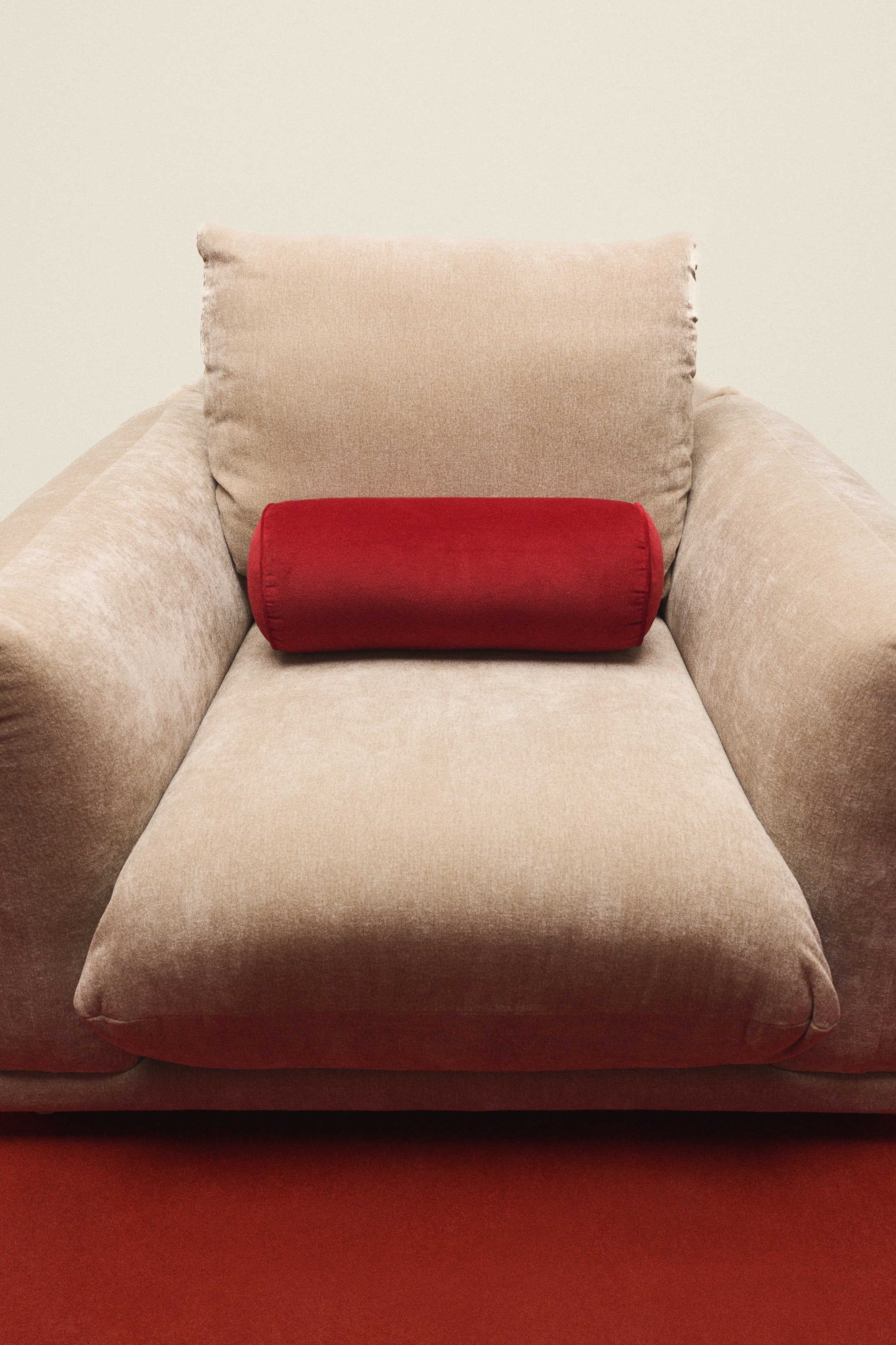 View larger image: A beige velvet armchair is shown from the front, with a red cylindrical bolster cushion resting horizontally against its back cushion.