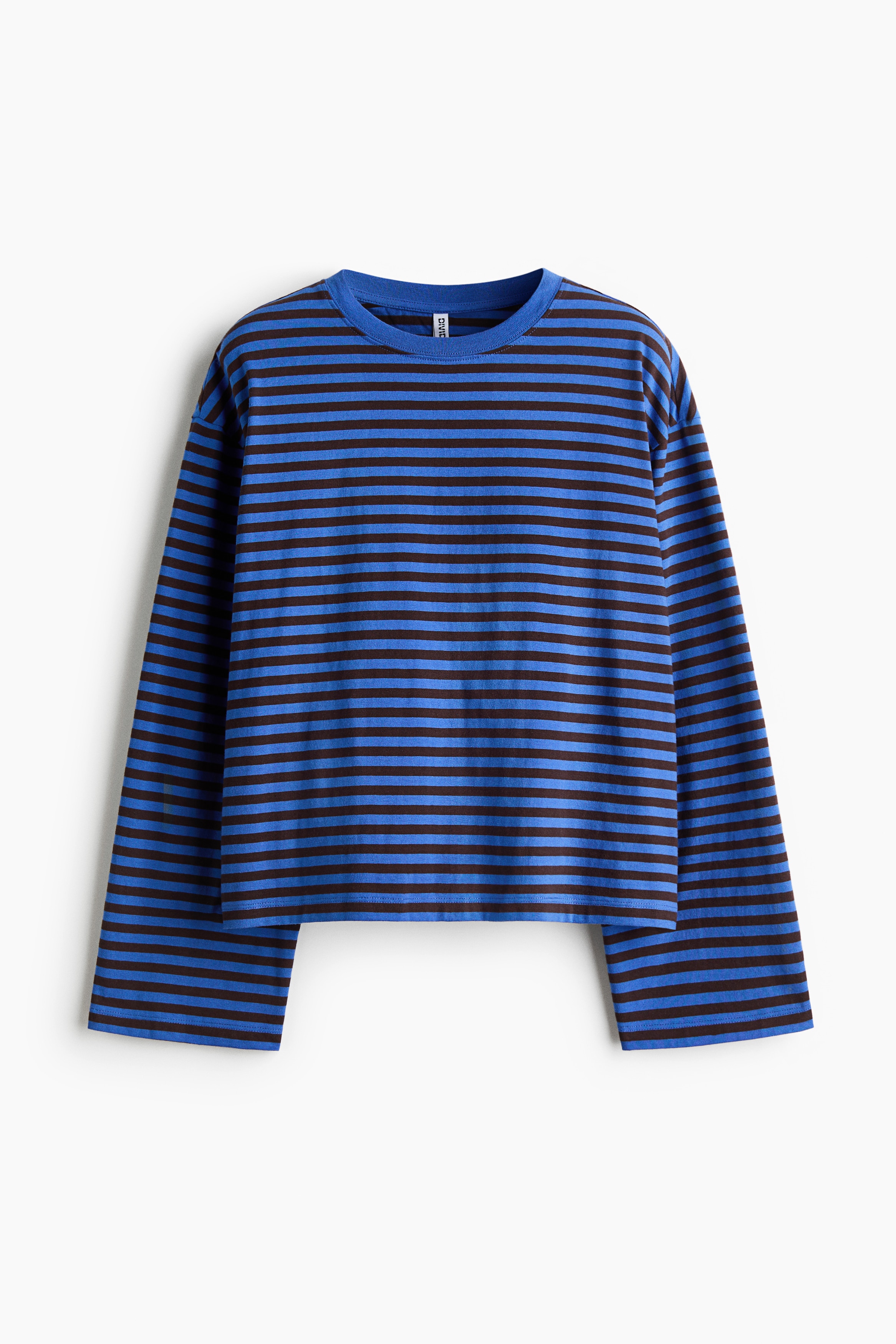 A loose-fit, long-sleeved top in a blue and brown striped pattern is being worn by the model, who is holding one sleeve. The top features a round neckline and dropped shoulders and is styled with knee-length denim shorts and tall black boots.