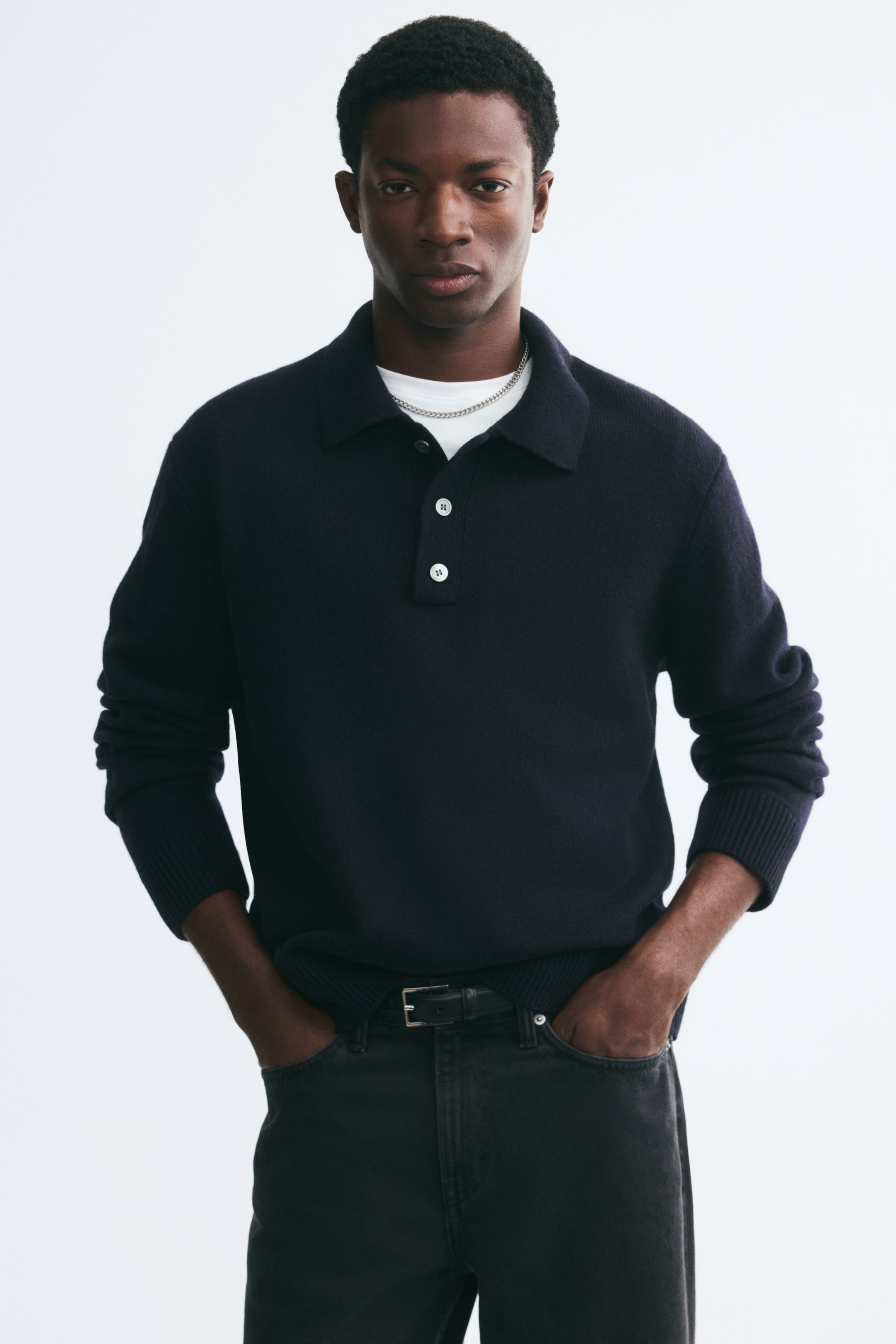 View larger image: A dark navy, fine-knit polo shirt is worn by the model, featuring a classic collar, a button placket with contrasting white buttons, and ribbed cuffs. The shirt is layered over a white T-shirt and styled with dark jeans.