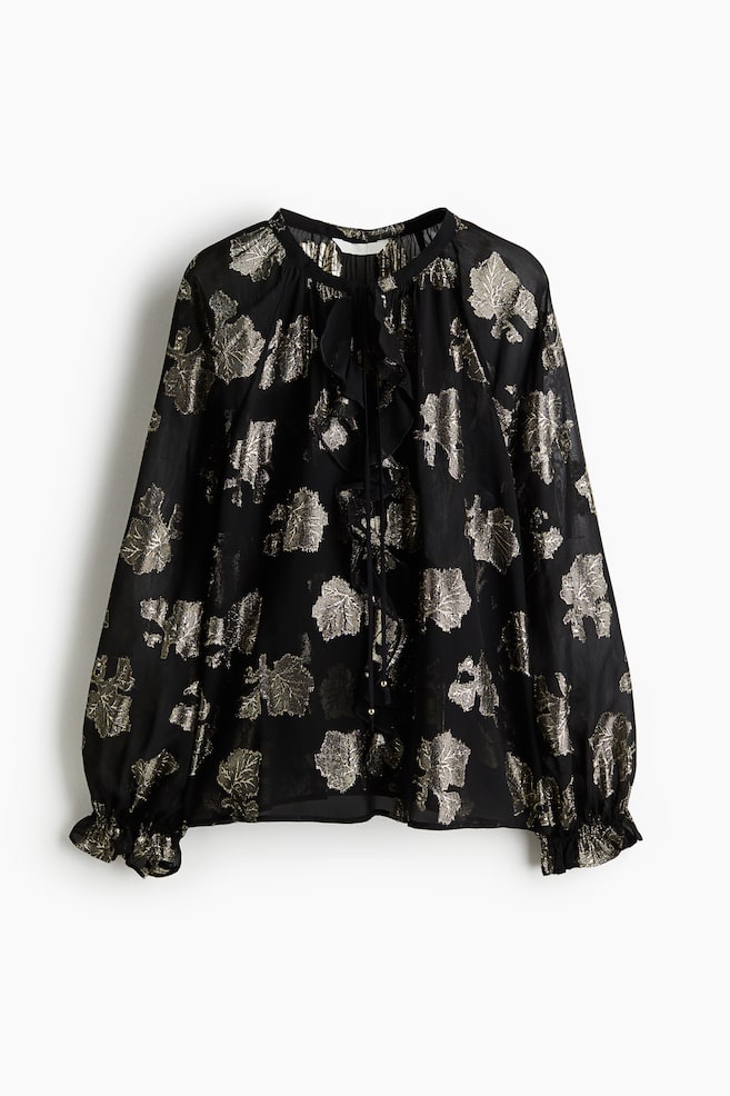 A black sheer blouse with a metallic gold leaf pattern is being displayed from the FRONT. It is designed with a gathered round neckline, a central ruffle placket with tie closures, and voluminous long sleeves are ending in ruffled cuffs. - 1