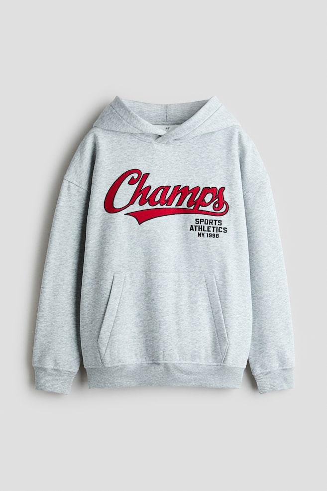 Se muestra una sudadera con capucha de mezcla gris claro vista de frente, con un grÃ¡fico cursivo rojo "Champs" en el pecho y texto negro que dice "SPORTS ATHLETICS NY 1998". La prenda de corte holgado presenta hombros caÃ­dos, capucha cruzada en el delantero, bolsillo tipo canguro y ribetes acanalados en los puÃ±os y el bajo. - 1