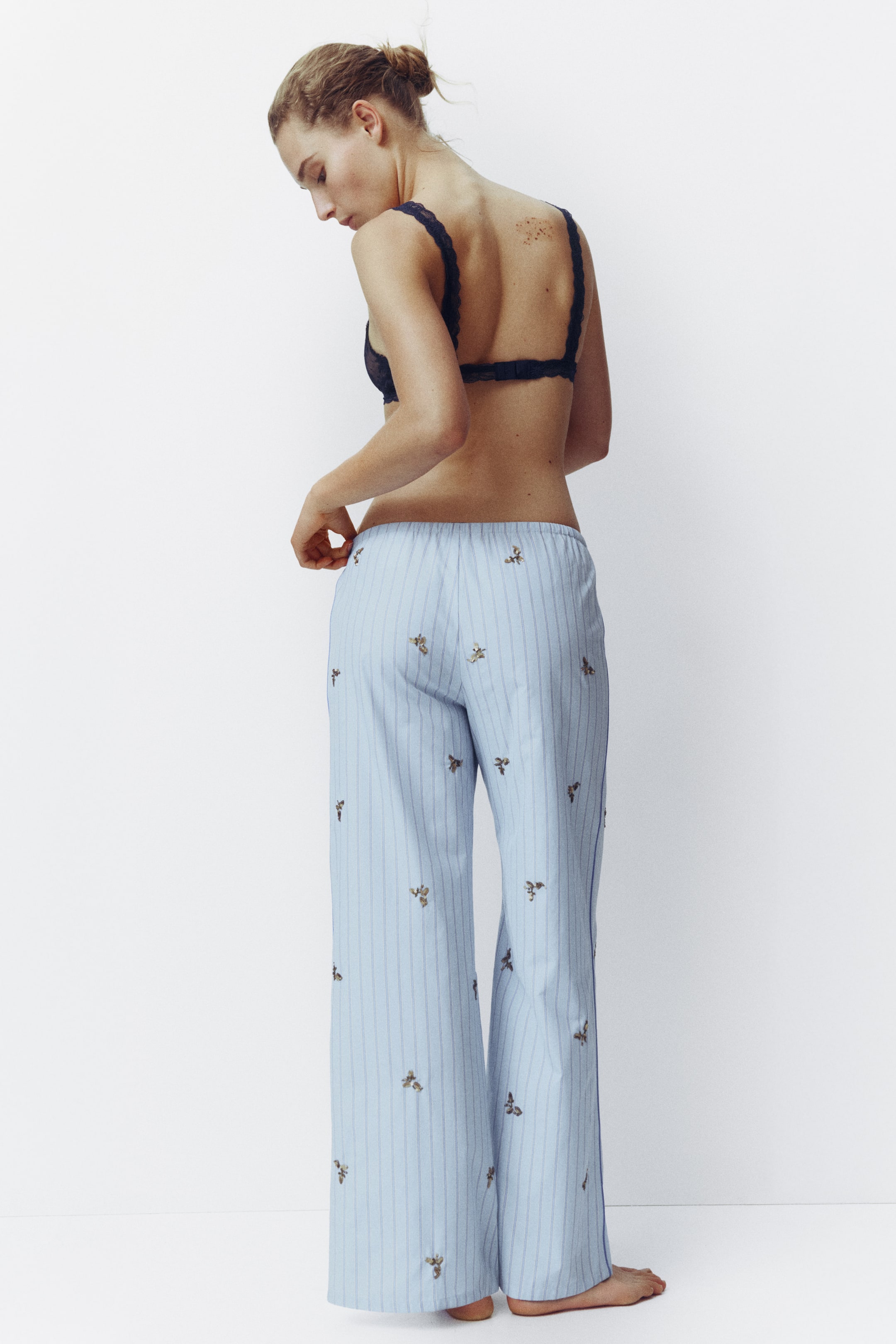 View larger image: The model is showcasing pyjama bottoms crafted from a light blue cotton weave adorned with vertical stripes, beads, and sequins. She is gently touching the waistband. These bottoms boast a high, elasticated waist and straight legs, highlighted by accent-colour piping along the sides.