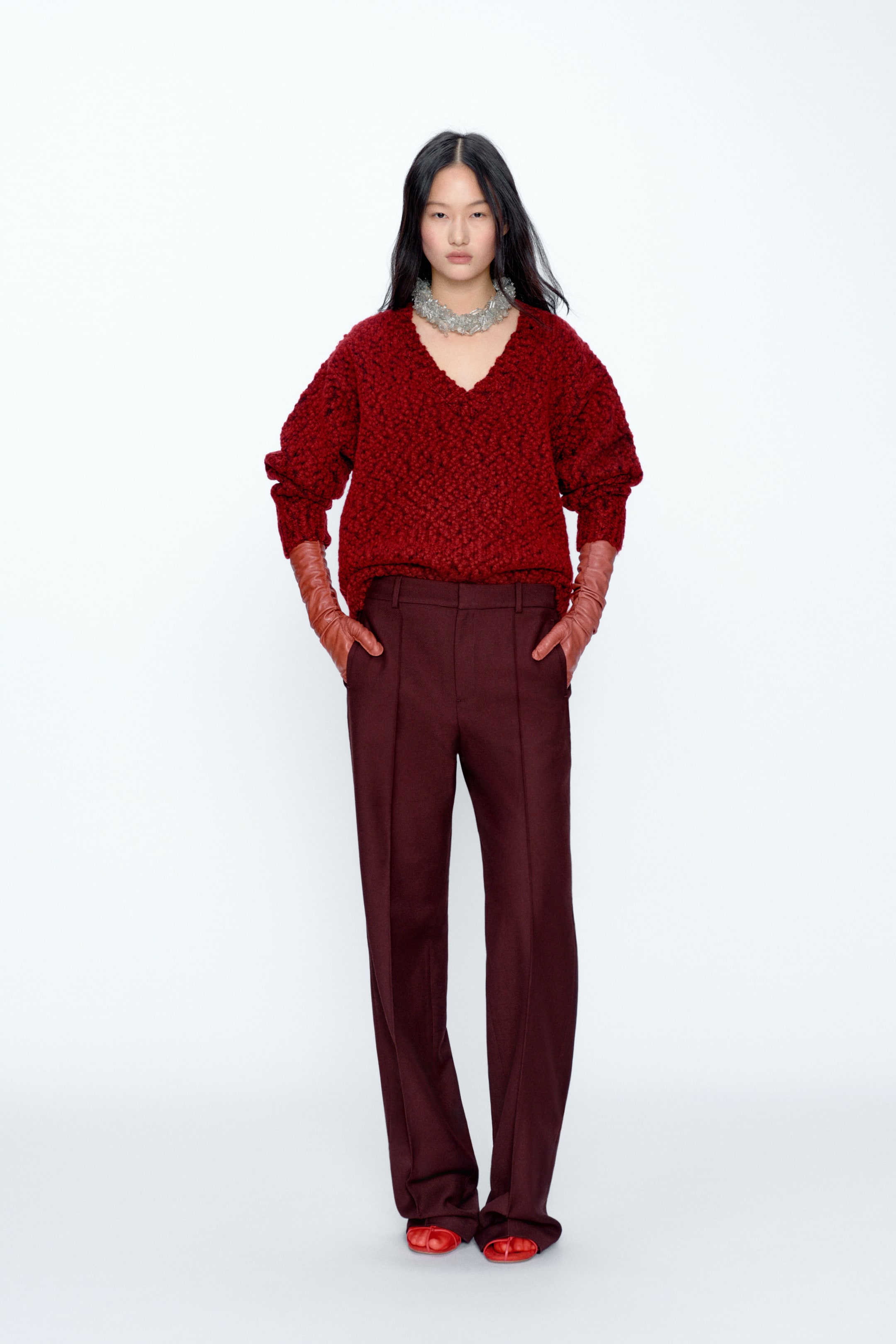 View larger image: A red textured V-neck sweater with puffed sleeves is styled with burgundy straight-leg trousers, shown from the front. Long brown gloves extend from the sweater sleeves, red open-toe flats complete the look, and a chunky silver necklace adorns the neckline.