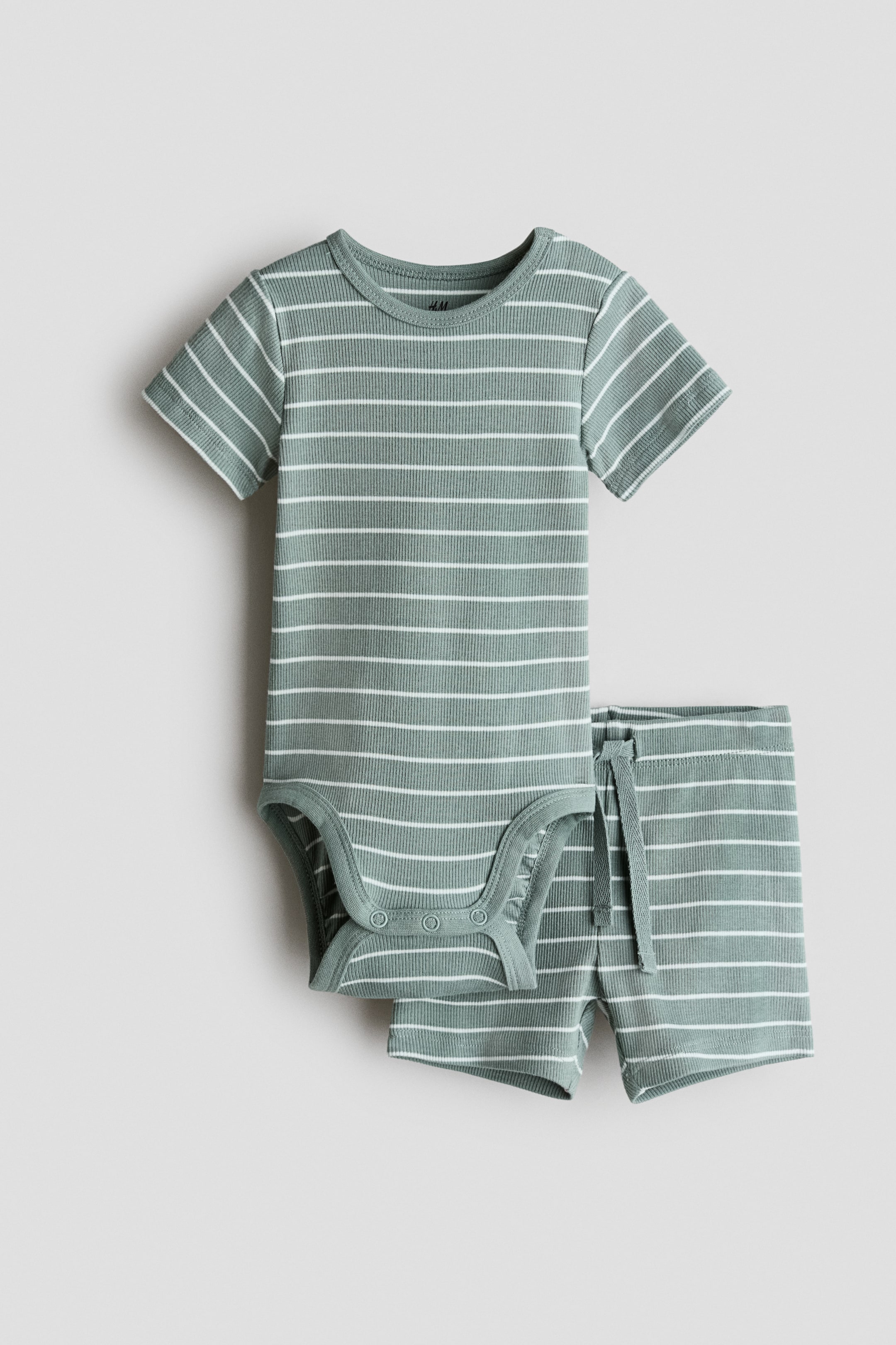 Grotere afbeelding bekijken: De tweedelige set bestaat uit een bodysuit met korte mouwen en een short, beide gemaakt van geribbelde katoenen jersey met saliegroene en witte horizontale strepen, gepresenteerd aan de voorkant. De bodysuit heeft een ronde halslijn en drie drukknopen bij het kruis, terwijl de short een bedekte elastische tailleband met een zichtbare trekkoord toont.