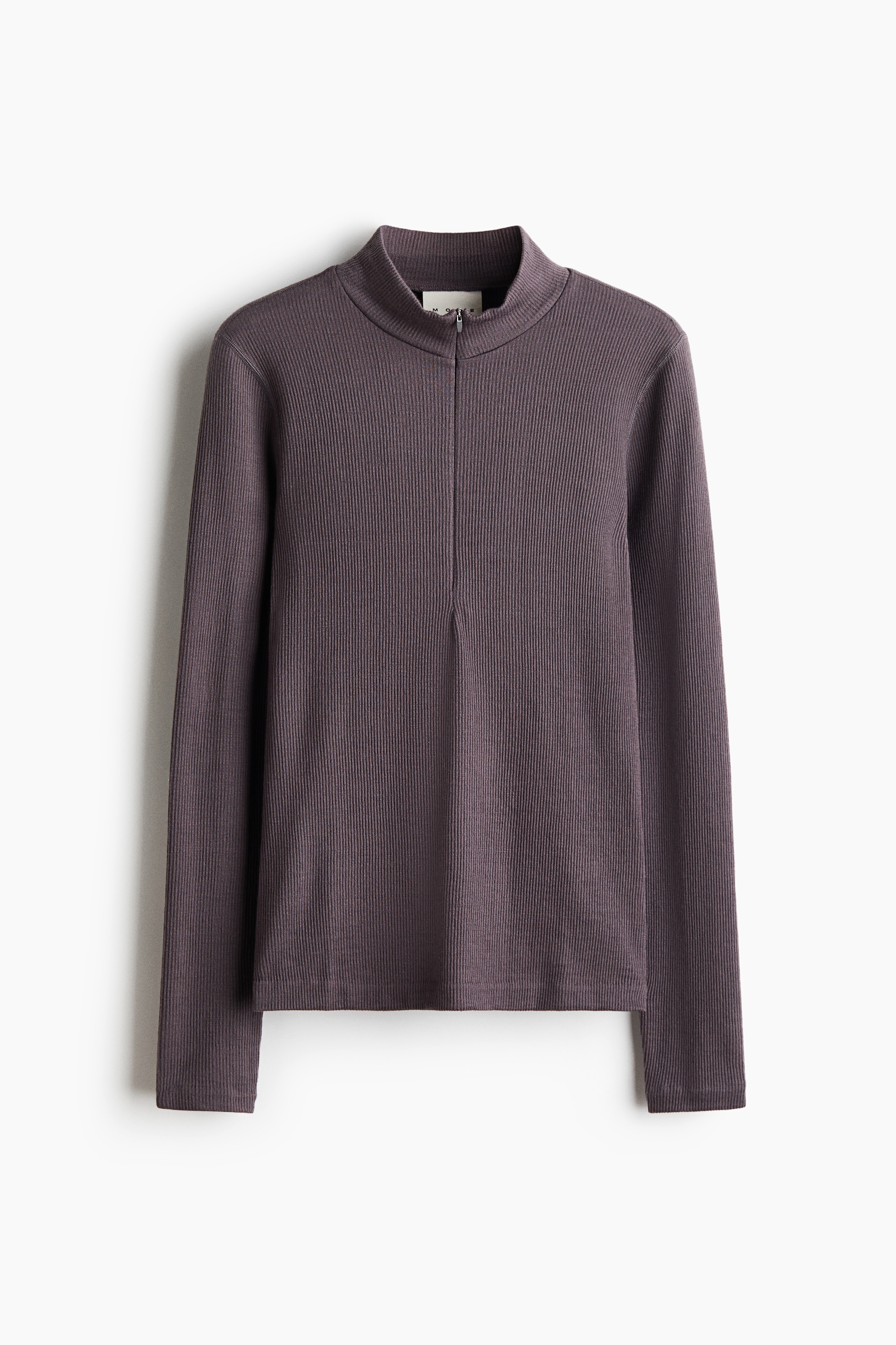 A dark purple outfit consisting of a ribbed, long-sleeved mock-neck top with a front zipper and matching slim-fit trousers is being worn by the model. The top is tucked into the high-waisted trousers, which feature side zipper pockets and a double-button closure.