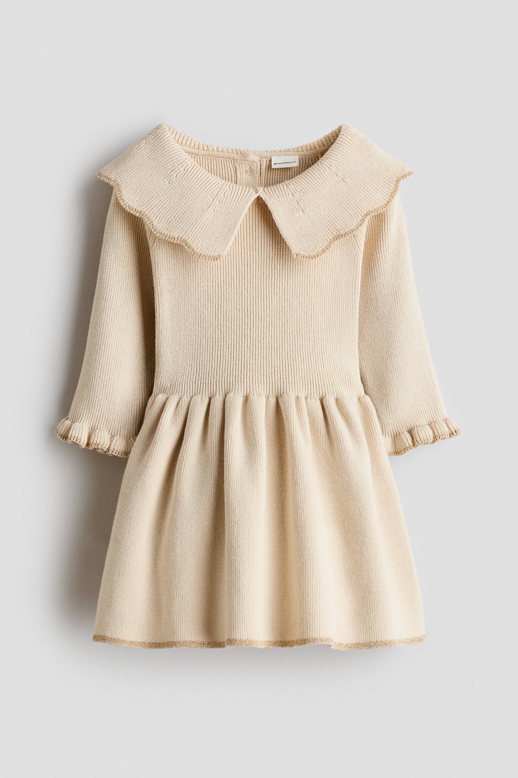 Agrandir l'image: Une robe en maille beige clair est présentée de face, dotée d’un grand col festonné aux détails en maille subtils et d’une bordure délicate. La robe met en valeur un corsage en maille côtelée et des manches longues, avec des poignets volantés et une taille froncée menant à une jupe évasée en maille côtelée, le tout rehaussé d’une bordure assortie au col, aux poignets et à l’ourlet.
