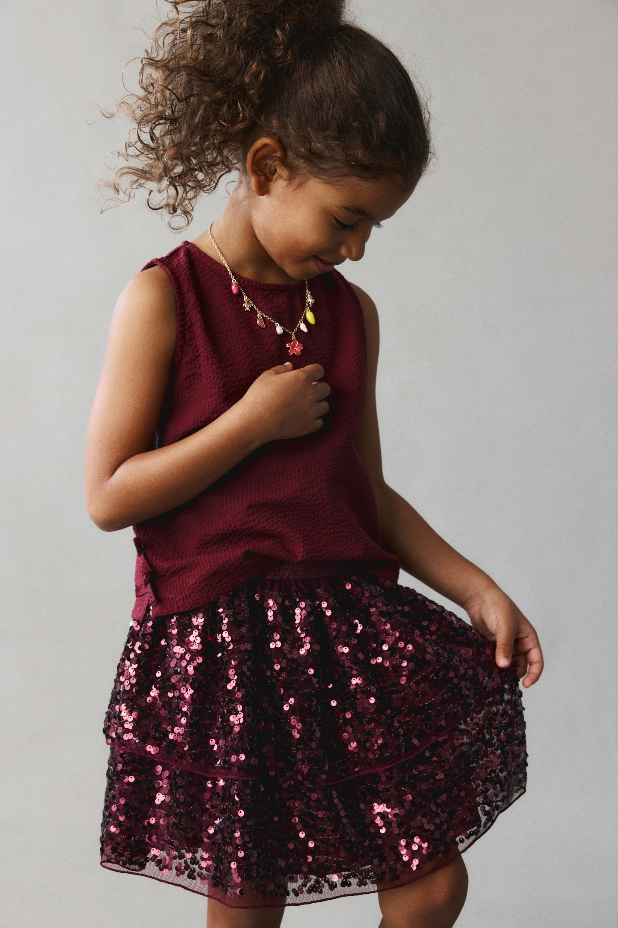 View larger image: A burgundy tulle skirt covered in shimmering sequins is shown from the front, featuring a tiered, flared design. The skirt is paired with a matching textured sleeveless top, highlighting its glittery elasticated waistband and lightweight, layered structure.