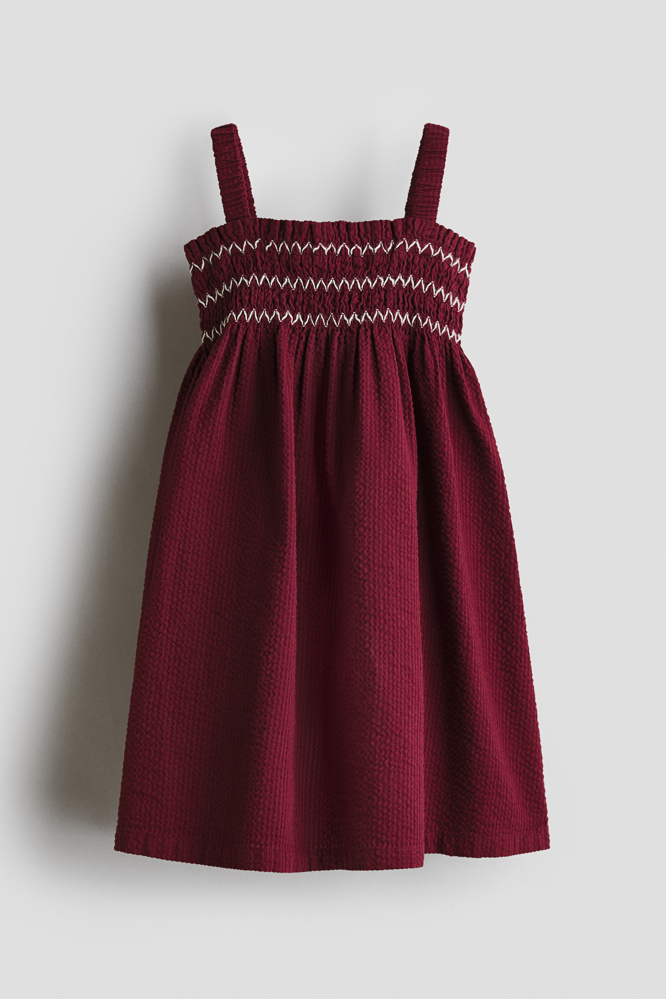 A dark red seersucker dress is shown from the front, featuring a smocked bodice with white zig-zag embroidery, a square neckline with a small frill trim, elasticated shoulder straps, and a flared skirt.