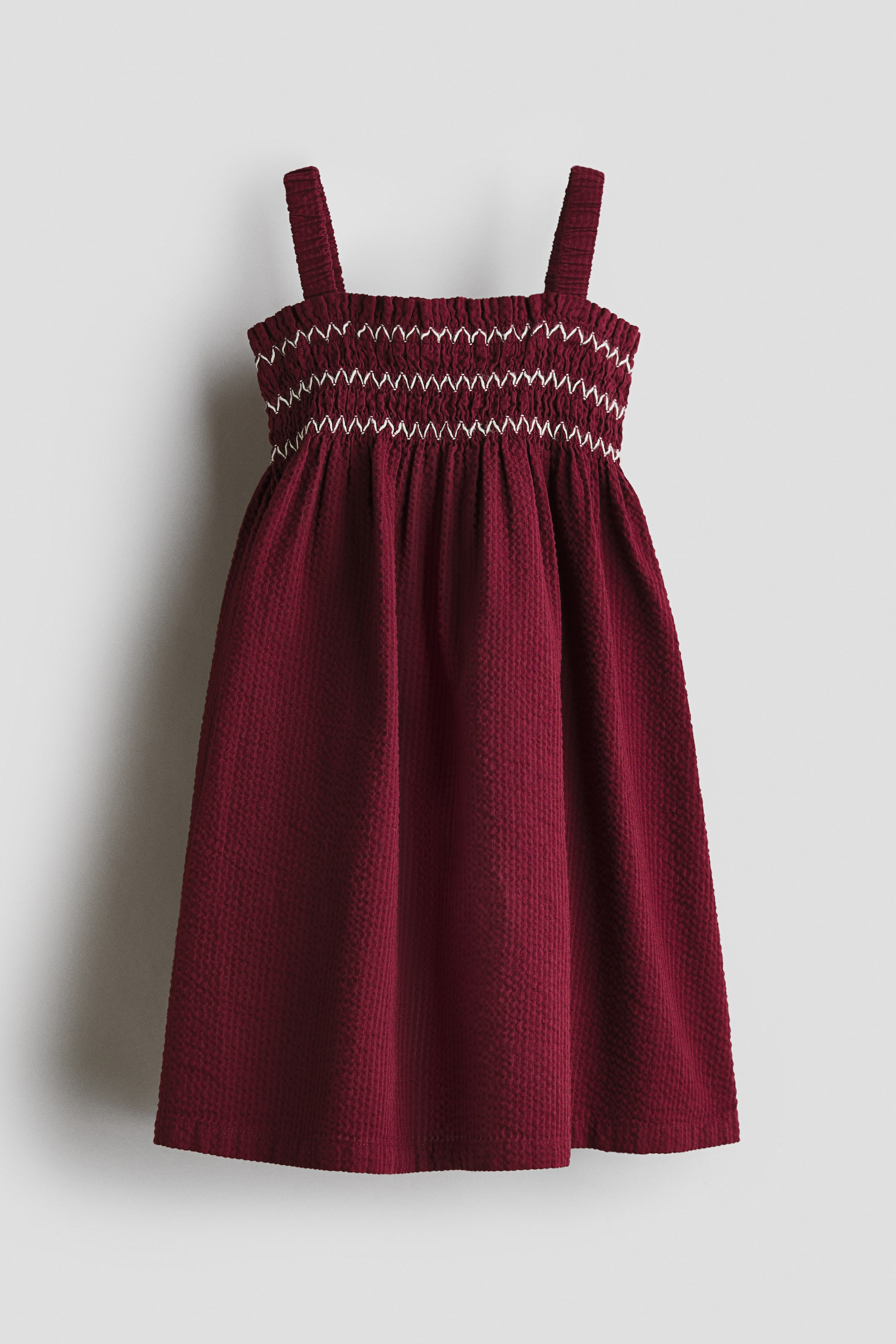 View larger image: A burgundy seersucker dress is shown from the front, featuring a smocked bodice detailed with white zigzag embroidery and broad shoulder straps. The dress flows into a gathered, flared skirt made from a finely textured, crinkled fabric.