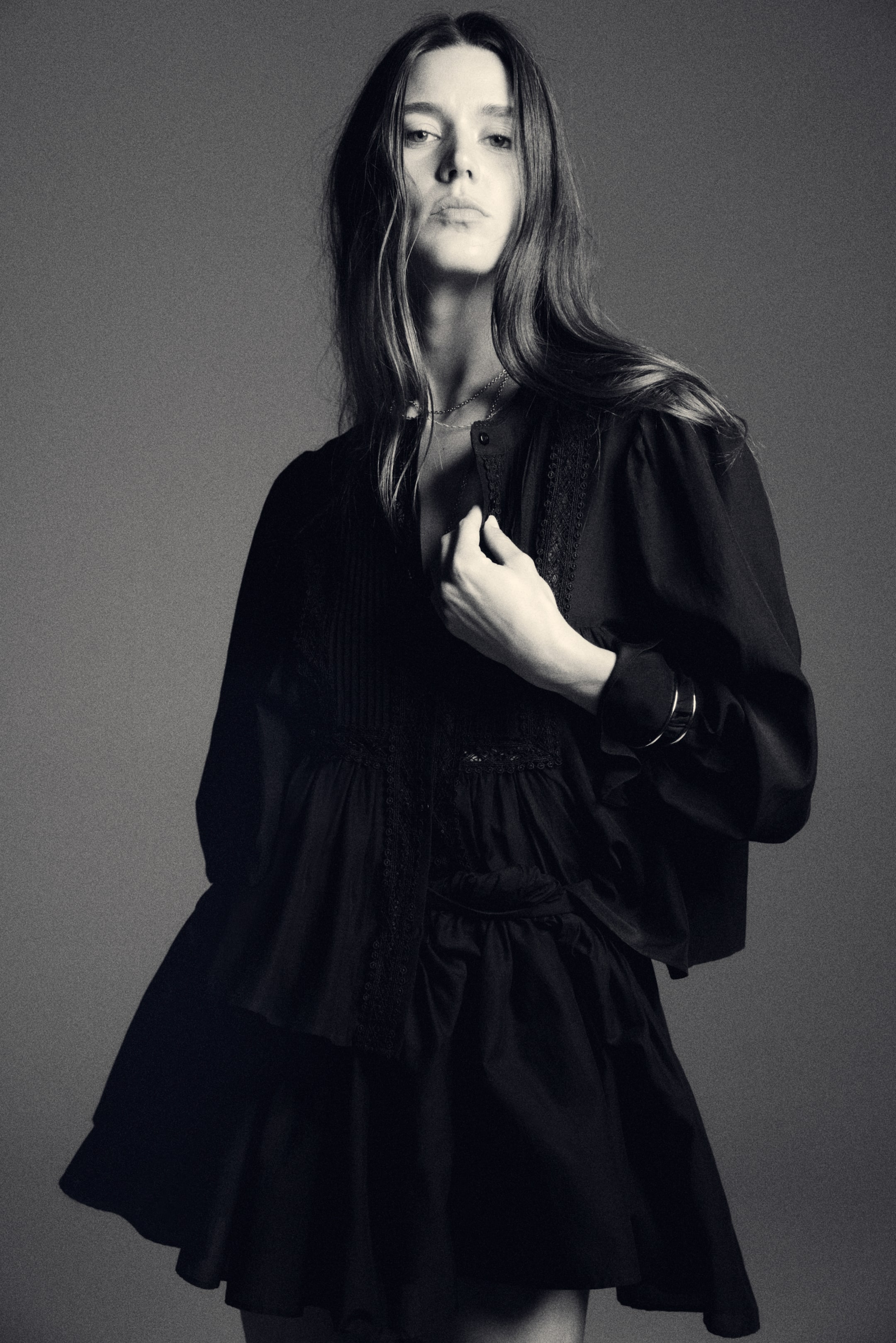 View larger image: A black collarless blouse is featuring a partial button placket, pin-tuck detailing, and lace trim on the bodice, with long gathered sleeves. The blouse is flowing into a peplum hem, layering over a tiered black mini skirt that is flaring out.