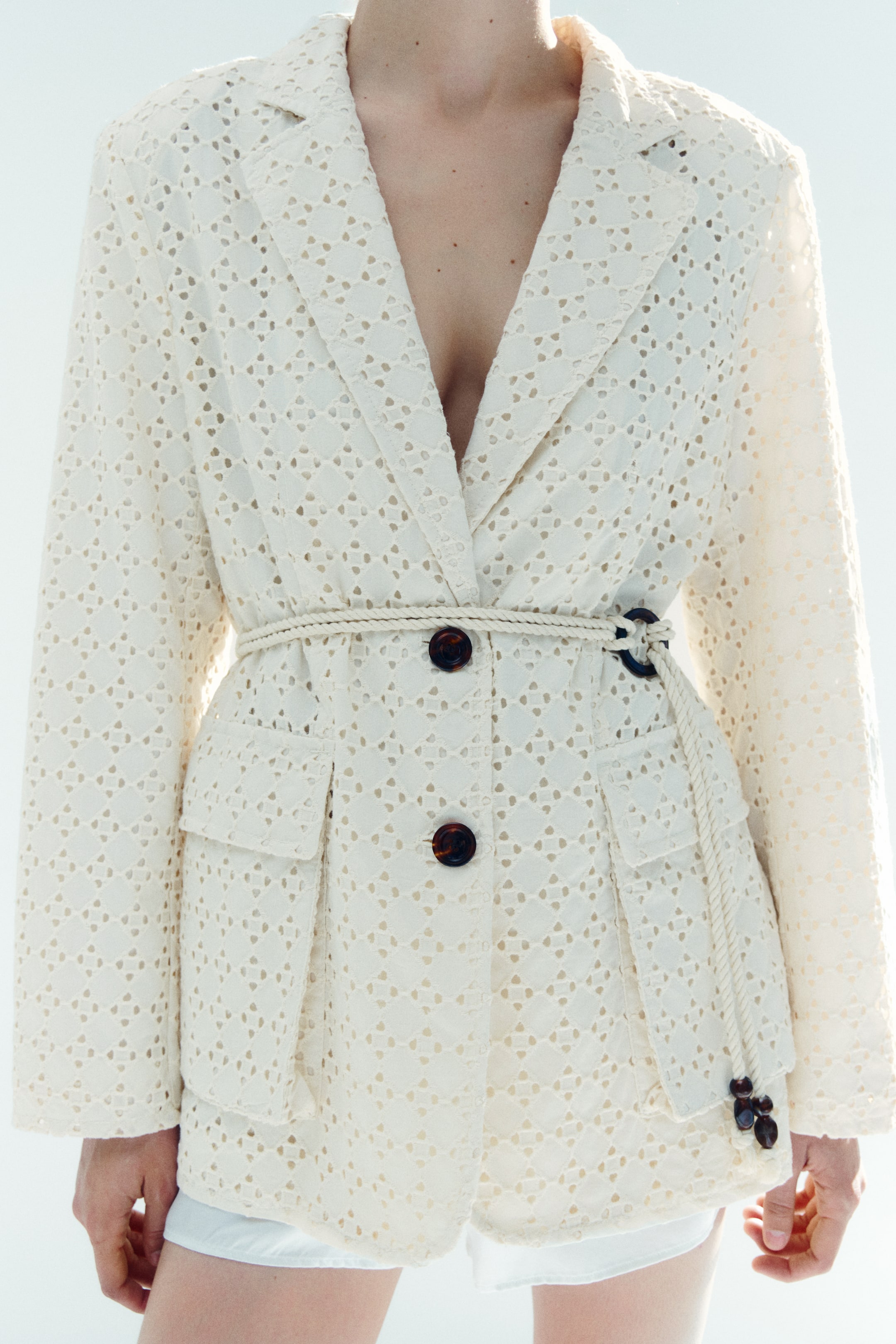 View larger image: A cream eyelet blazer is facing forward, featuring notch lapels and a perforated diamond pattern. It showcases two brown tortoise-shell buttons and two flap patch pockets, with a cream rope belt cinching the waist, tied with a dark ring and beads on the right side.