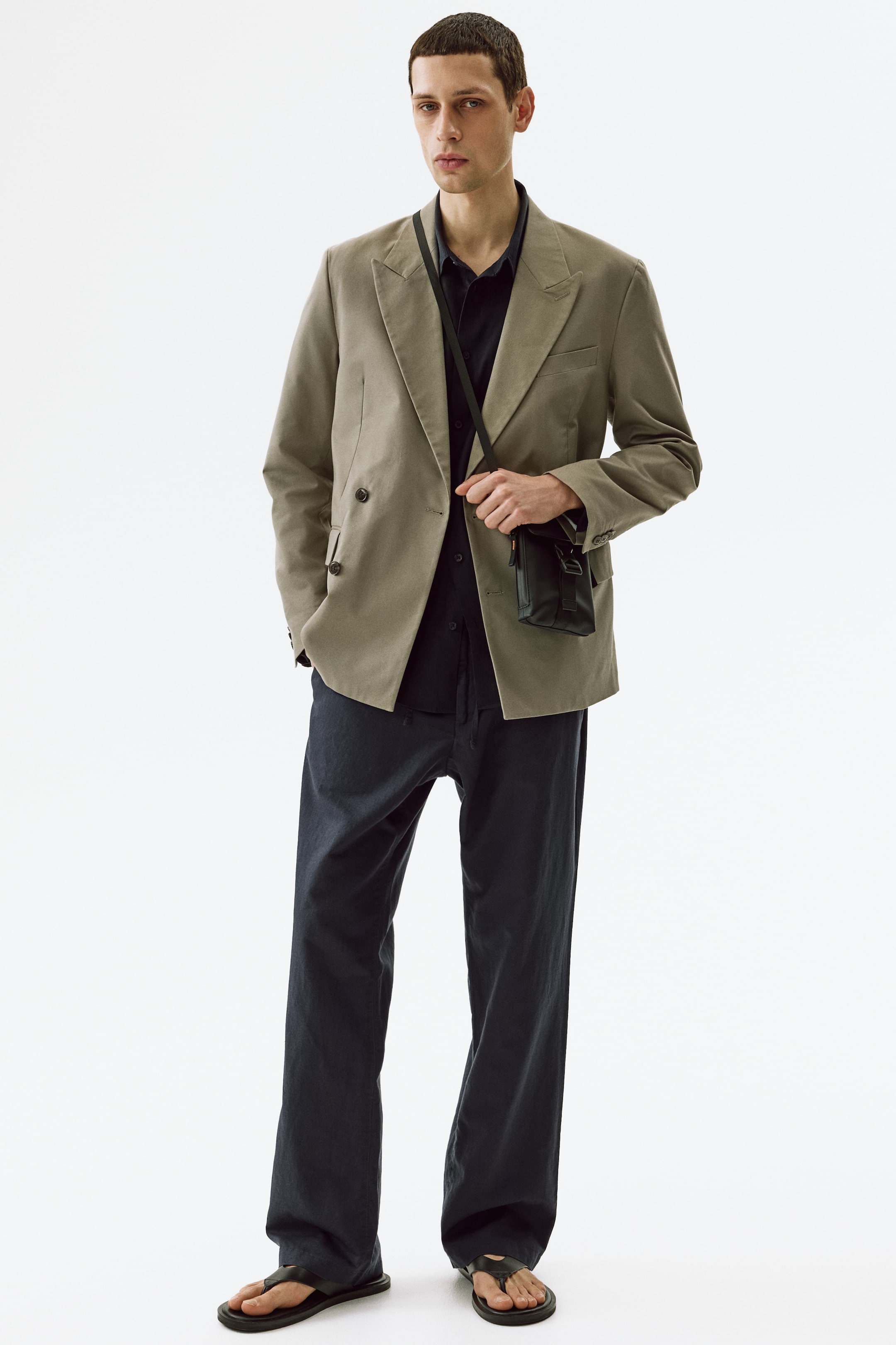 View larger image: A light olive double-breasted blazer features wide notch lapels, two visible rows of dark buttons, and flap pockets on the lower front. It is styled open over a dark shirt, with a black crossbody bag draped across its front.