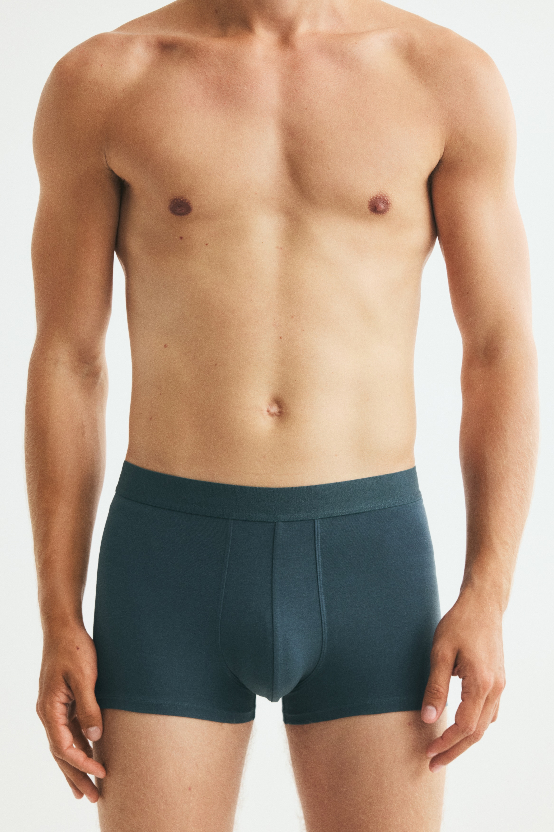 Vier Paar Boxershorts werden in einem Fächerstapel präsentiert, mit elastischen Bundabschlüssen. Sie erscheinen in den Farben Dunkelgrün, Schwarz und Türkis, wobei das unterste türkise Paar ein strukturiertes Vorderteil aufweist.