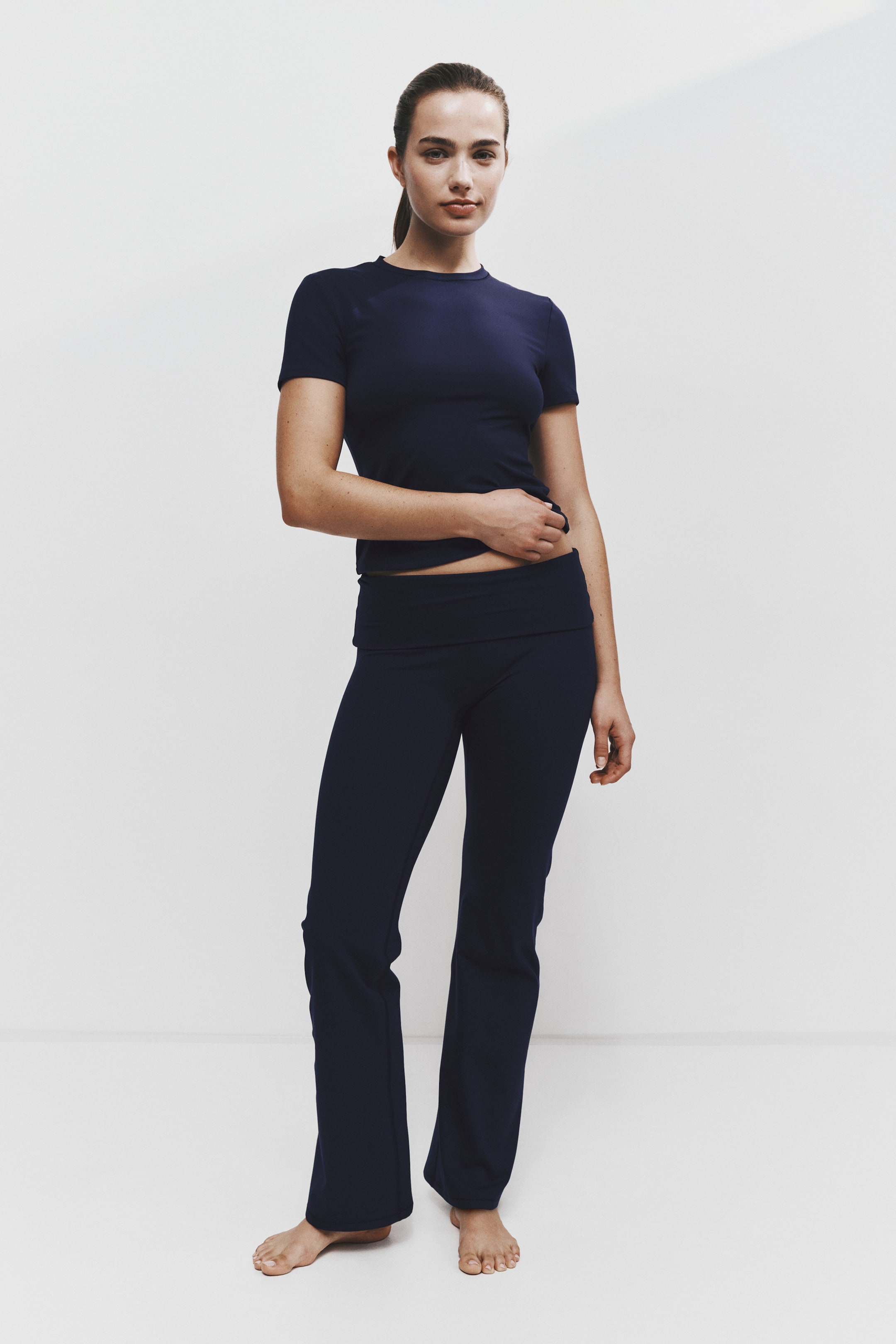 Visualizza l'immagine ingrandita: Un completo activewear abbinato blu navy è indossato frontalmente, composto da una T-shirt slim fit con scollo tondo e maniche corte e pantaloni coordinati. I pantaloni presentano un’ampia fascia in vita ripiegata e una silhouette a gamba svasata.