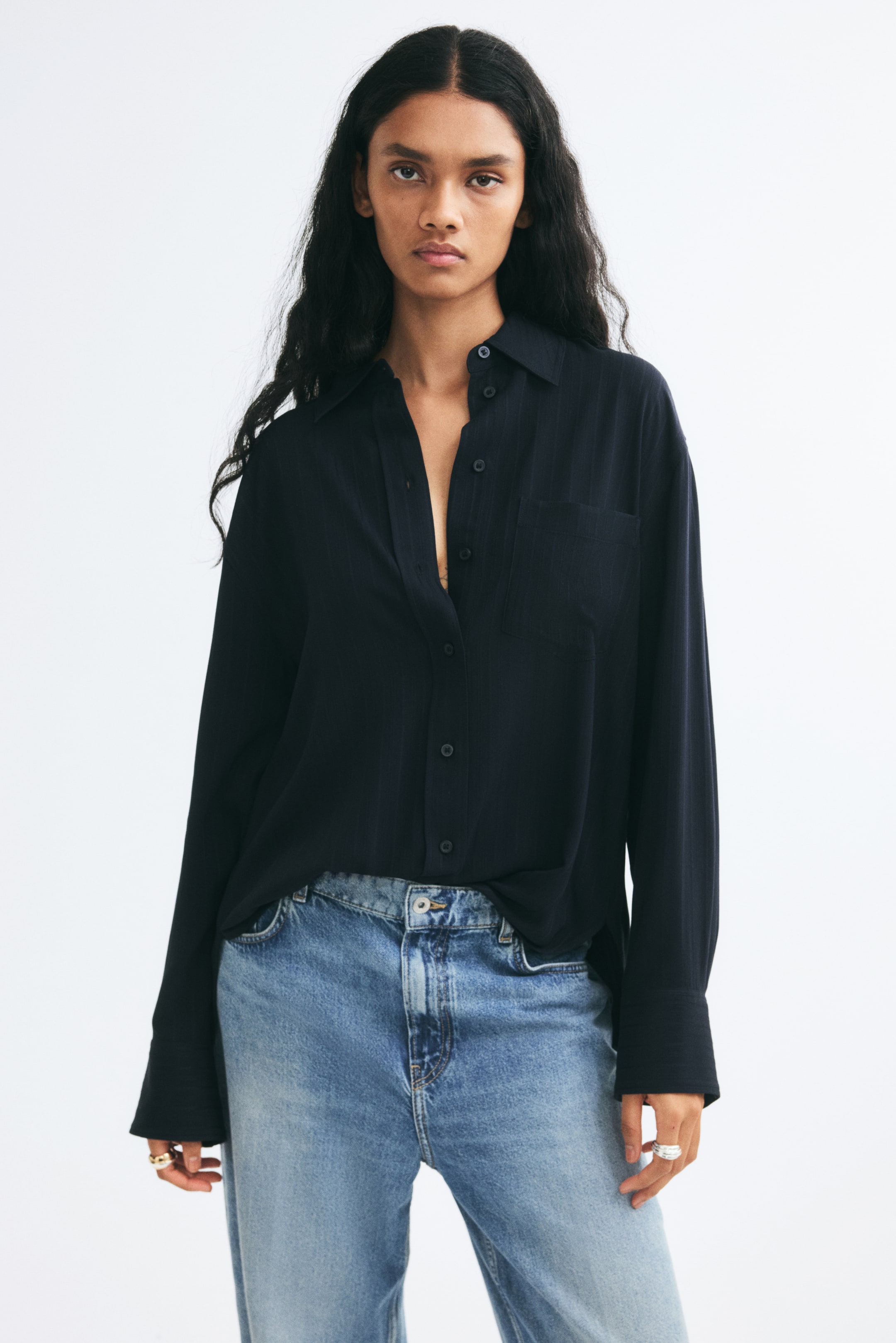 Visa större bild: En svart oversized skjorta i vävd struktur bärs av modellen och är delvis instoppad i ljusblå jeans. Skjortan har en krage, knäppning framtill, en enkel bröstficka med påsydd ficka samt långa ärmar med sänkta axlar och vida manschetter.