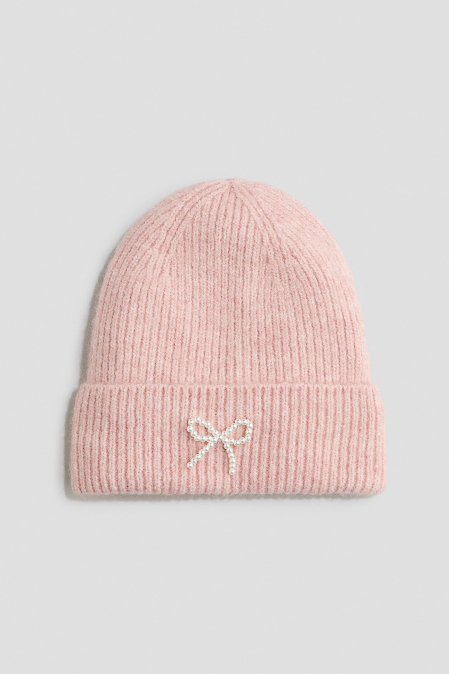Un berretto a maglia a coste, rosa tenue, è rivolto frontalmente e mette in risalto un ampio risvolto ripiegato impreziosito da un piccolo fiocco composto da perle bianche. - 1