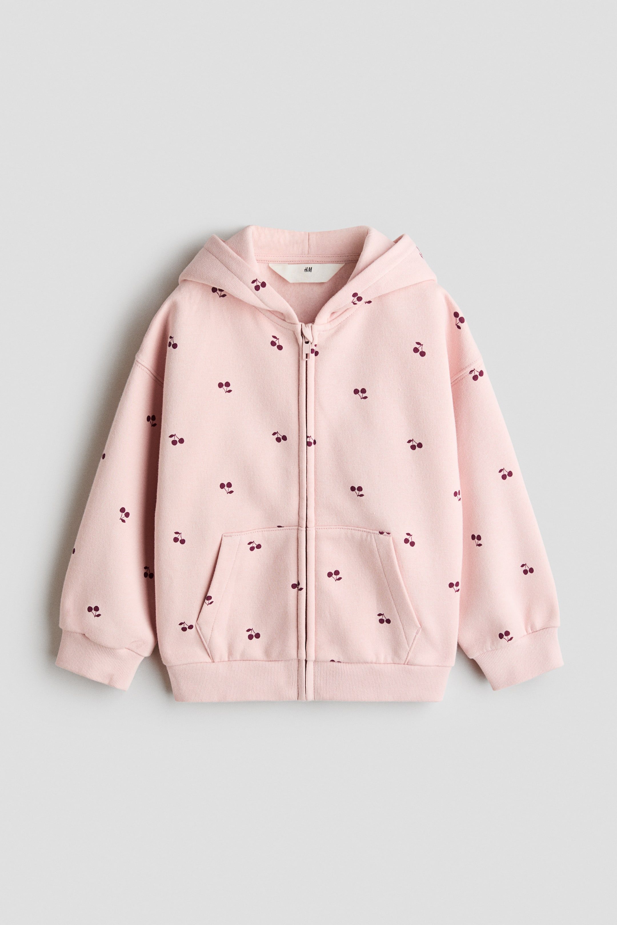 View larger image: A pink full-zip hoodie is laid flat and facing forward, showcasing an all-over pattern of small dark red cherries. It features a full front zipper, an attached hood, two front patch pockets, ribbed cuffs, and a ribbed hem.