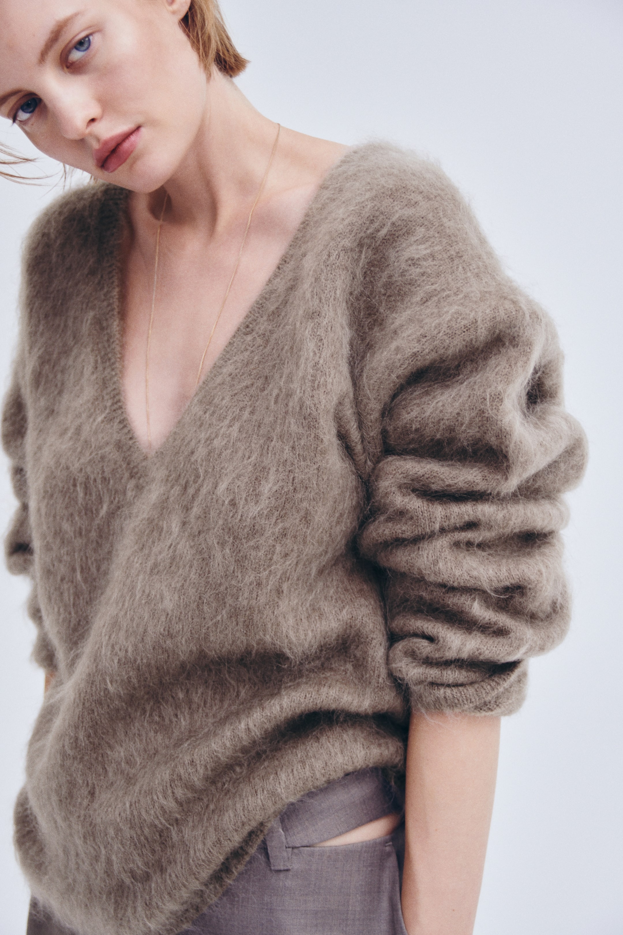 Visualizza l'immagine ingrandita: Una maglia oversize in maglia spazzolata color tortora, con profondo scollo a V e spalle scese, indossata dalla modella. Le maniche lunghe e materiche sono arrotolate sulle braccia, e il maglione è abbinato a pantaloni grigi.