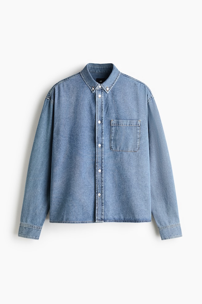 A light blue denim shirt is shown from the front, featuring a button-down collar, a full-length button placket with white buttons, and a single patch pocket on the left chest. The shirt features dropped shoulders, long sleeves with buttoned cuffs, and a straight hemline. - 1