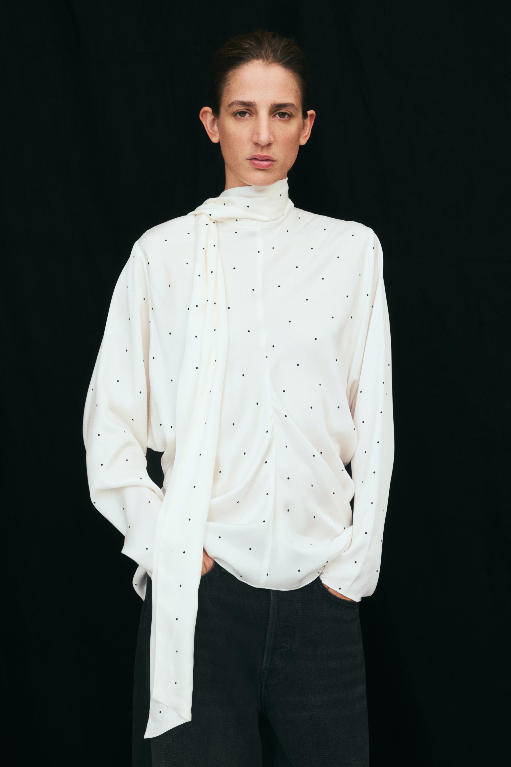 Ampliar la imagen: La modelo lleva una blusa blanca de lunares, con cuello alto tipo scarf y una larga cinta drapeada que cae al frente. La blusa tiene mangas largas y una costura central fruncida, y se combina con jeans negros.