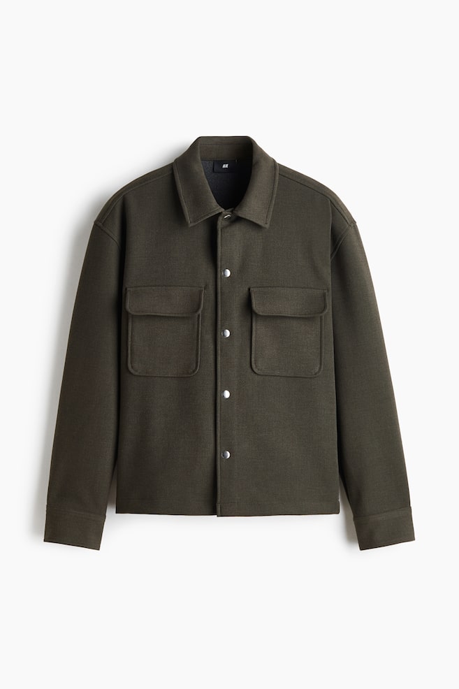 Se muestra de frente una sobrecamisa verde oliva oscuro, con un corte estructurado y cuadrado, cuello clásico y cierre frontal de broches plateados. Presenta dos grandes bolsillos de parche con tapa en el pecho y mangas largas con puños de acabado limpio. - 1