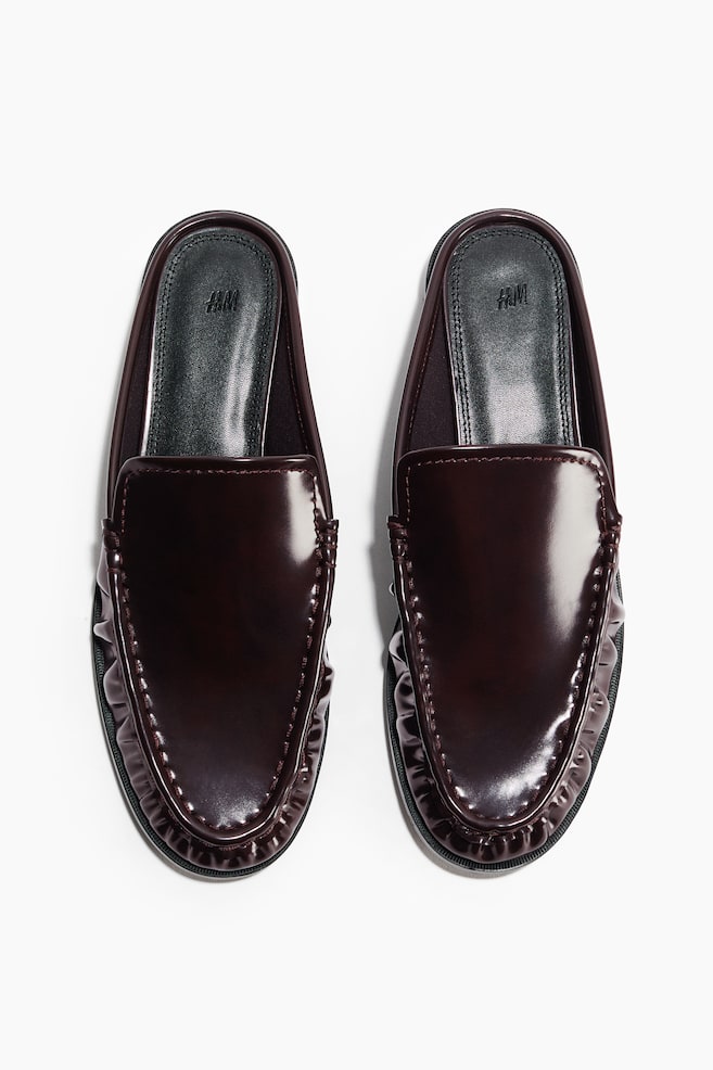 A pair of burgundy patent-finish backless loafers is showcased from a top-down perspective. They feature moccasin-style stitching and gathered detailing around the vamp. - 1