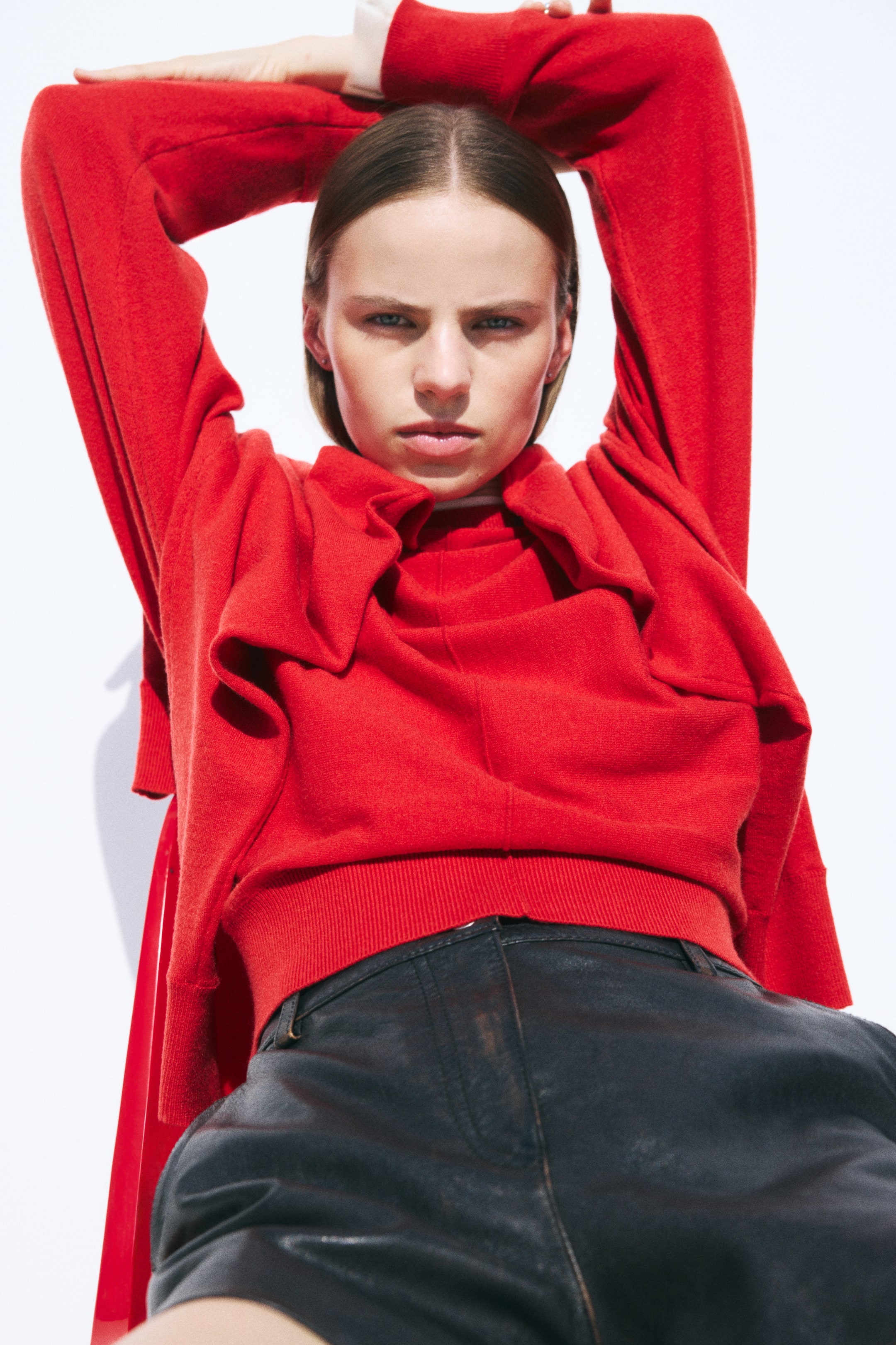 Größeres Bild anzeigen: Ein roter Strickpullover mit sichtbarer Frontnaht und geripptem Saum wird von dem Model getragen, das mit erhobenen Armen posiert. Ein passender roter Pullover liegt über den Schultern des Models und ist mit schwarzen Leder-Shorts kombiniert.