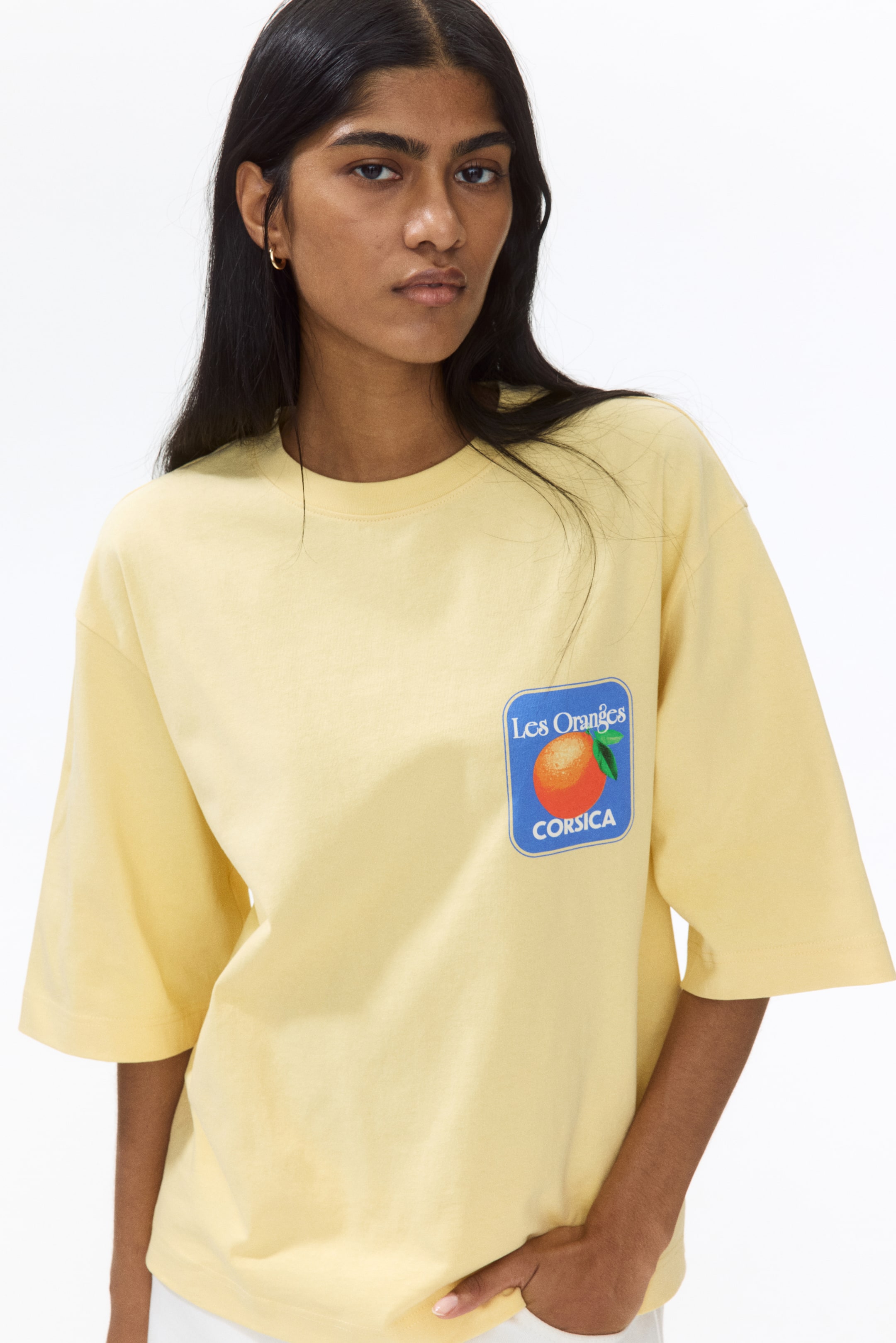 View larger image: A loose-fitting pale yellow t-shirt features a round neckline and short sleeves. On the left chest area, a square blue patch is visible, displaying an orange fruit and the text "Les Oranges CORSICA."