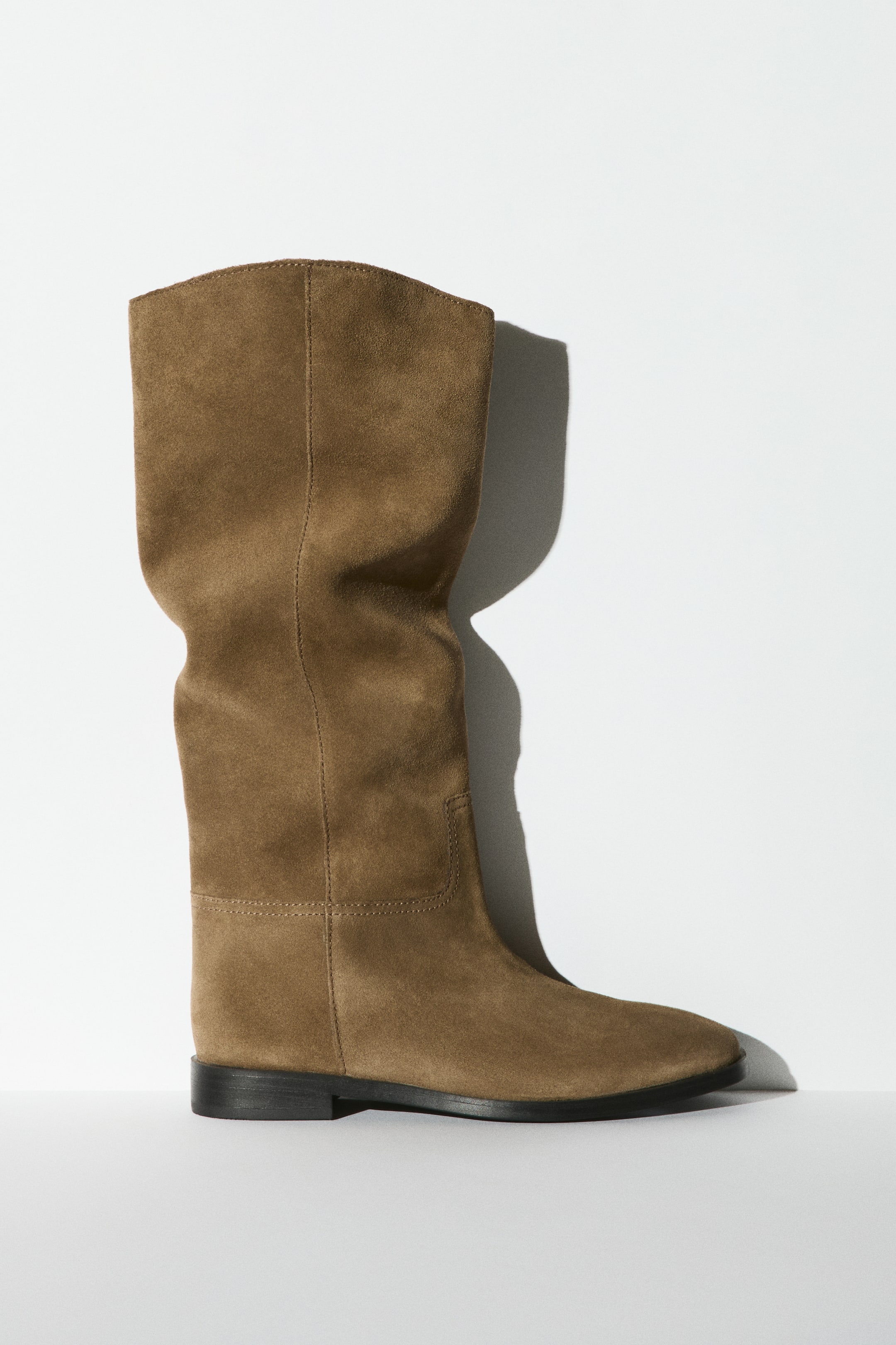 View larger image: A knee-high boot in soft brown suede is shown in side profile facing right, featuring a rounded toe, a flat black sole, and a relaxed, slightly slouchy shaft. The boot is constructed with visible tonal stitching detailing the foot and ankle area.