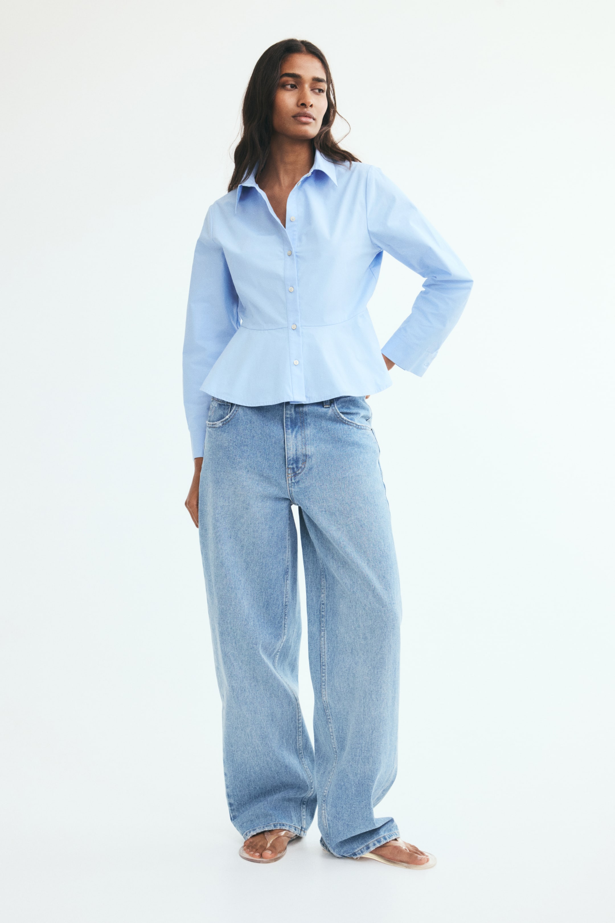 View larger image: A light blue cotton poplin shirt with a classic collar, front button closure, and a flared peplum hem is worn by the model. The long-sleeved shirt features buttoned cuffs and is paired with light-wash wide-leg jeans.