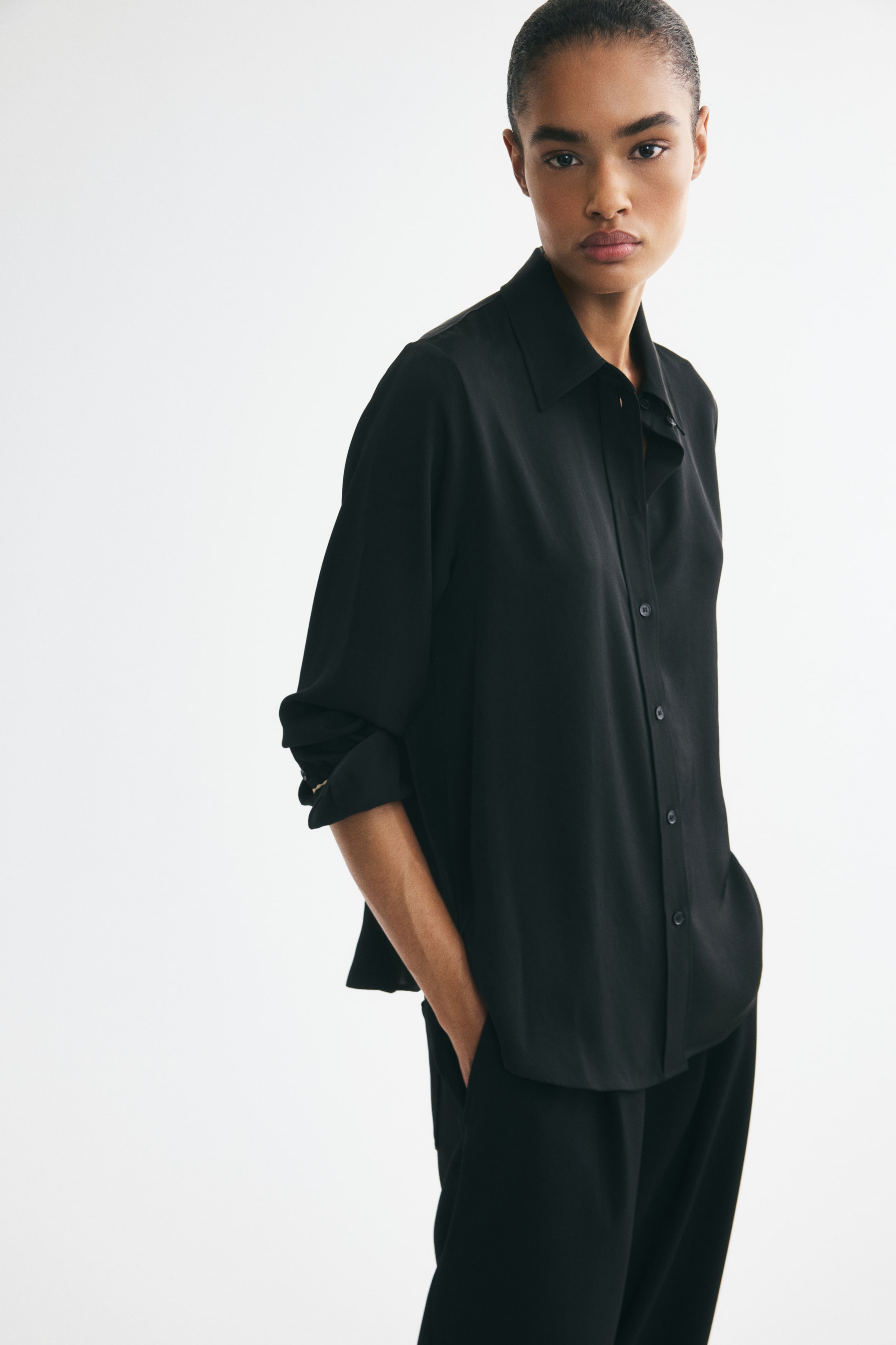 View larger image: The model is wearing a black woven shirt with a collar and buttons down the front. The shirt showcases a rounded hem, and the model is rolling up its long sleeves while placing one hand in a pocket.