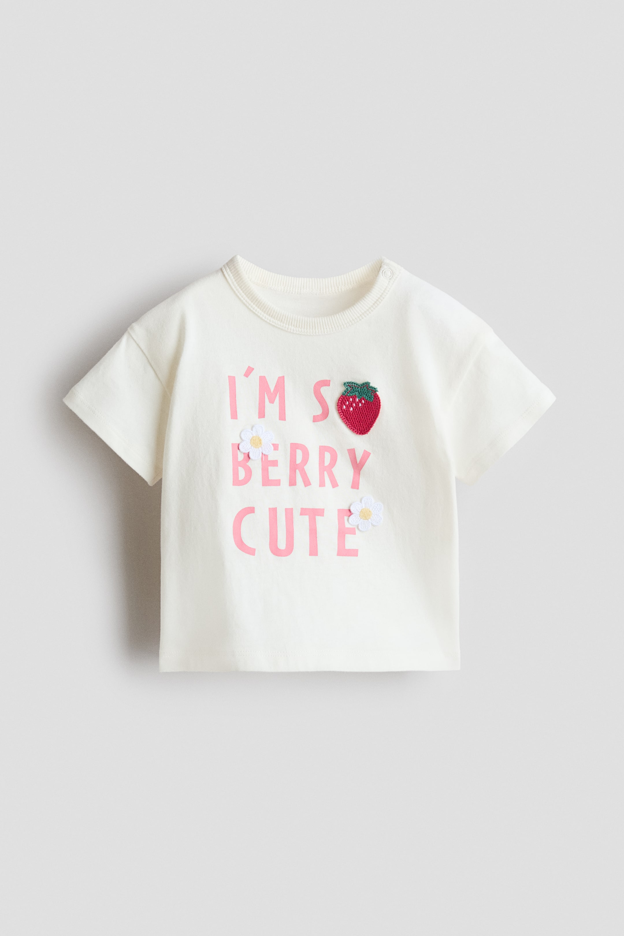View larger image: A cream T-shirt presents a ribbed crew neck with a snap button on the shoulder and short sleeves. The FRONT features "I'M SO BERRY CUTE" in pink lettering, along with a red strawberry applique and two white daisy appliques.