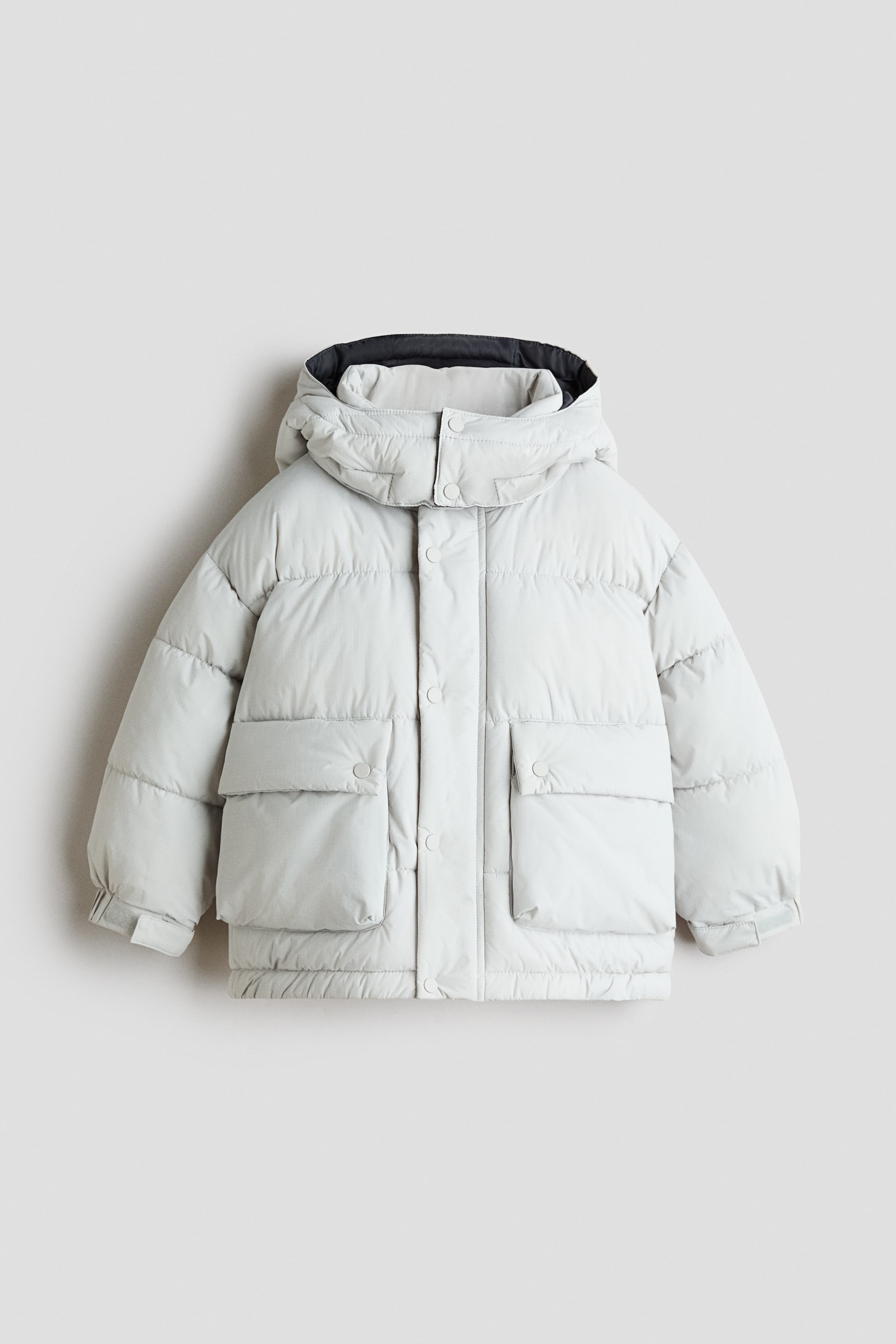 Ver imagen más grande: Una chaqueta acolchada gris claro vista de frente, con acolchado horizontal y capucha de cuello alto con forro interior oscuro. La chaqueta presenta un cierre frontal oculto con una solapa cortaviento de botones de presión, dos grandes bolsillos frontales con solapa y puños ajustables con tiras de cierre de gancho y bucle.