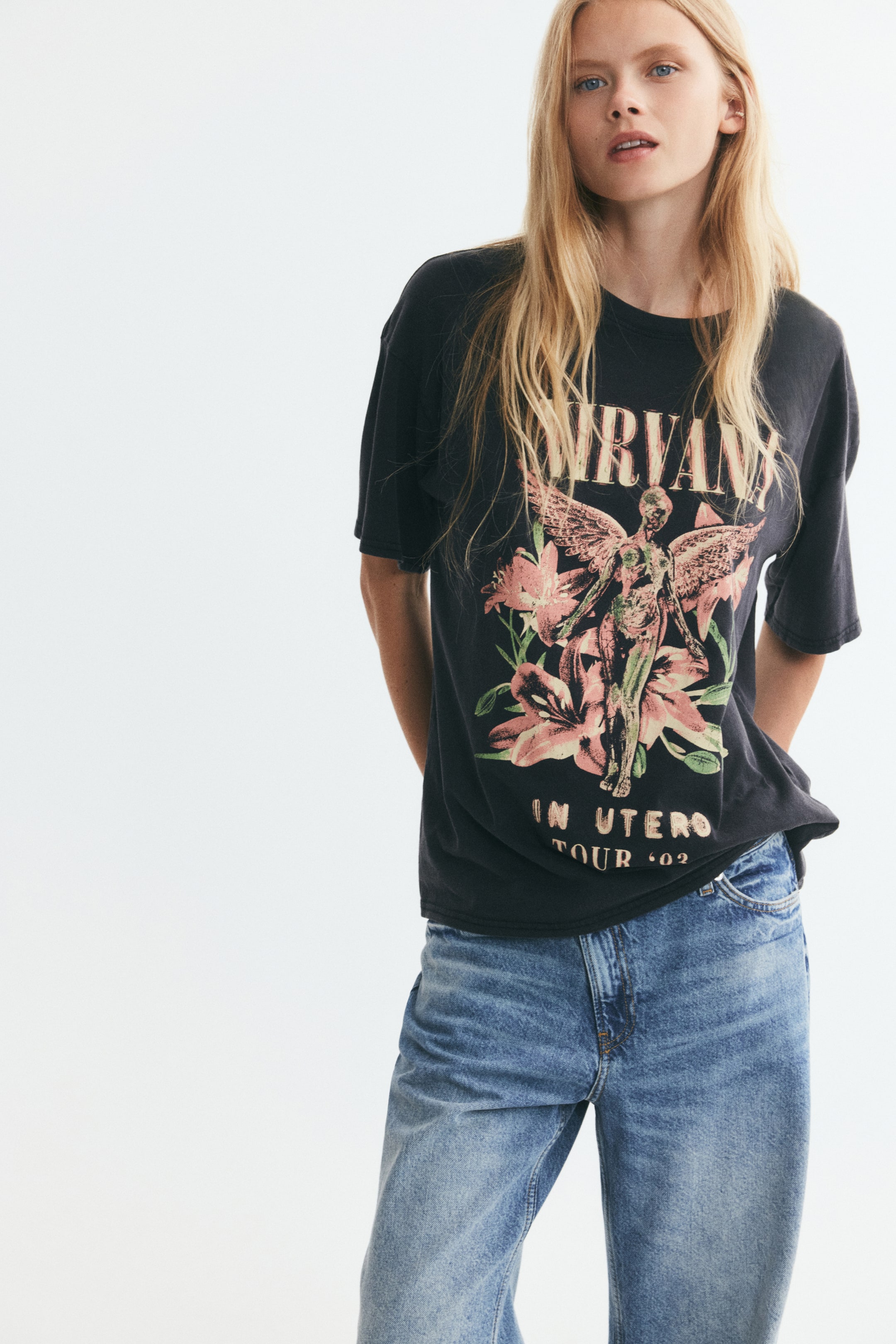 Προβολή μεγαλύτερης εικόνας: Ένα oversized, ξεβαμμένο μαύρο T-shirt με γραφικό print μιας φτερωτής φιγούρας και floral λεπτομέρειες φοριέται από το μοντέλο. Το T-shirt διαθέτει χαμηλωμένους ώμους και ribbed στρογγυλή λαιμόκοψη, και συνδυάζεται με μπλε denim τζιν.